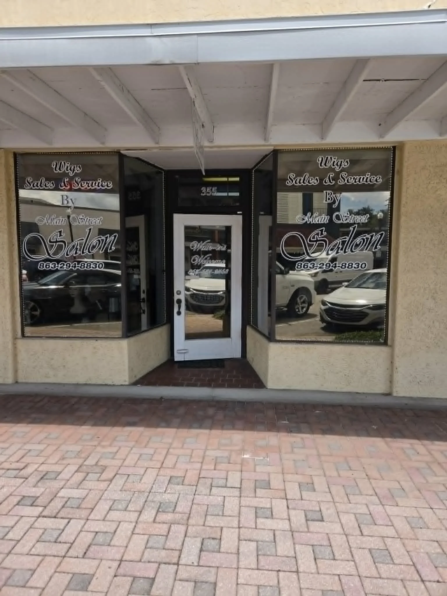 Storefront with glass doors and windows displaying the name 'Wigs Sales & Service by Main Street Salon', with a brick sidewalk in front and cars parked on the street reflected in the windows.