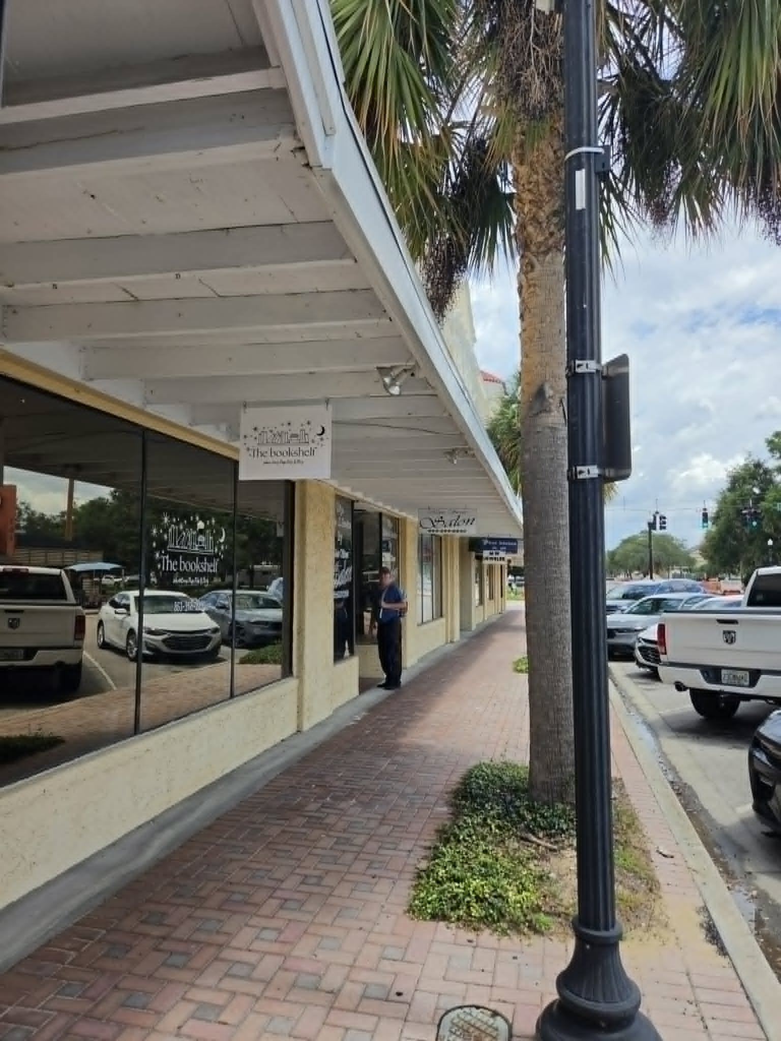 A storefront with large glass windows displaying the name ‘The bookshelf’ and a sidewalk with a brick pattern. There are parked cars along the street, a woman standing outside the store, a tall lamppost, and a palm tree nearby. The sky is partly cloudy.