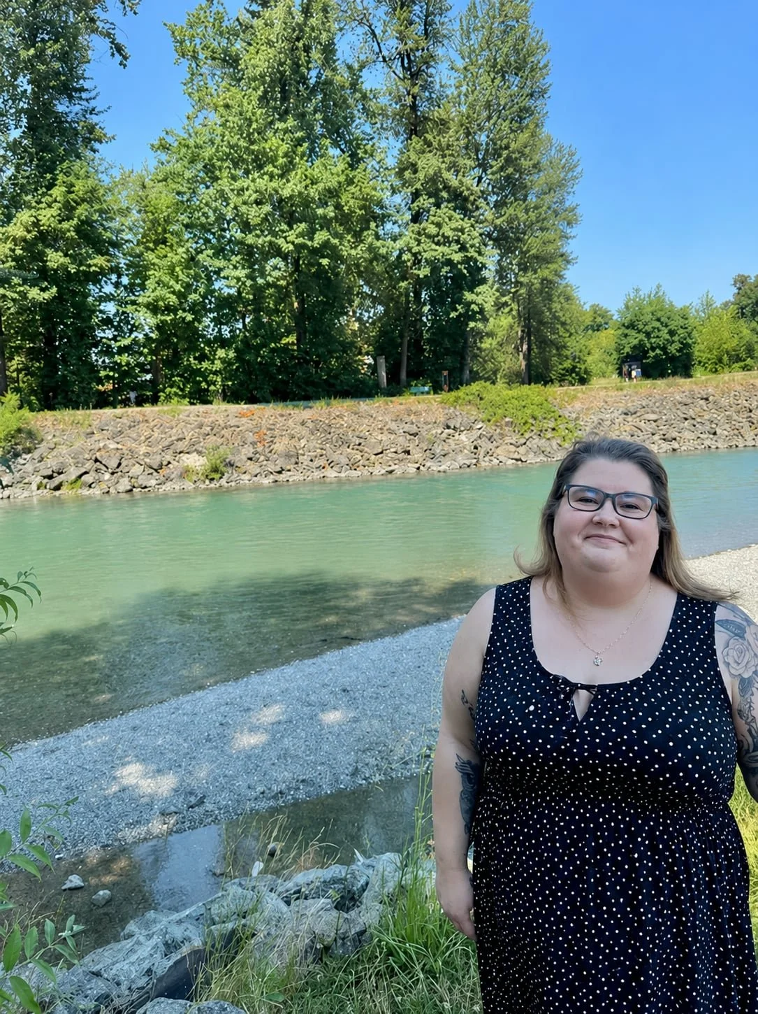 A woman with glasses and long hair, wearing a black dress with white polka dots, standing by a river on a sunny day with green trees and blue sky in the background.