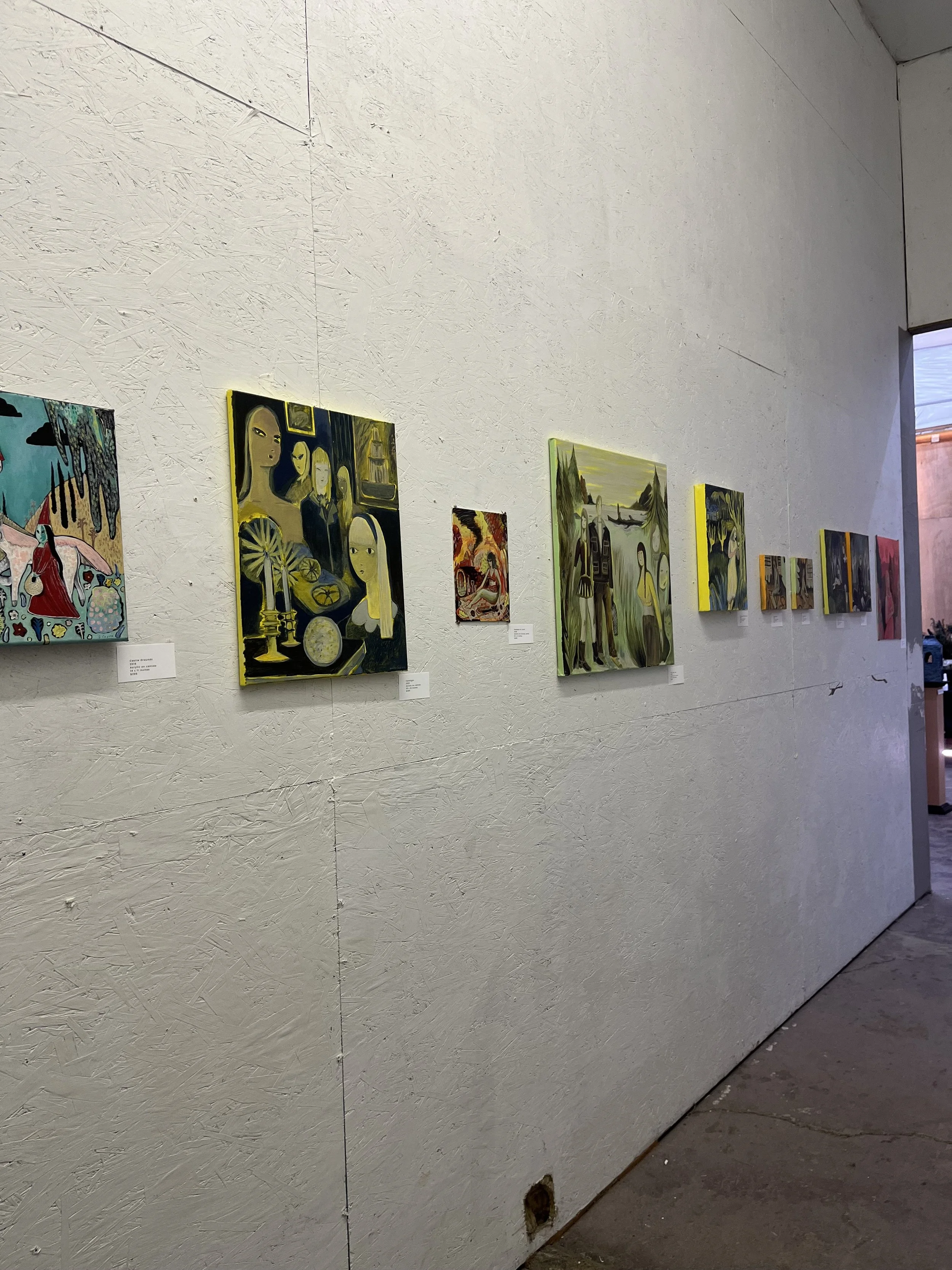 Gallery wall with multiple paintings of various styles and subjects, displayed on a textured white wall inside an art gallery.