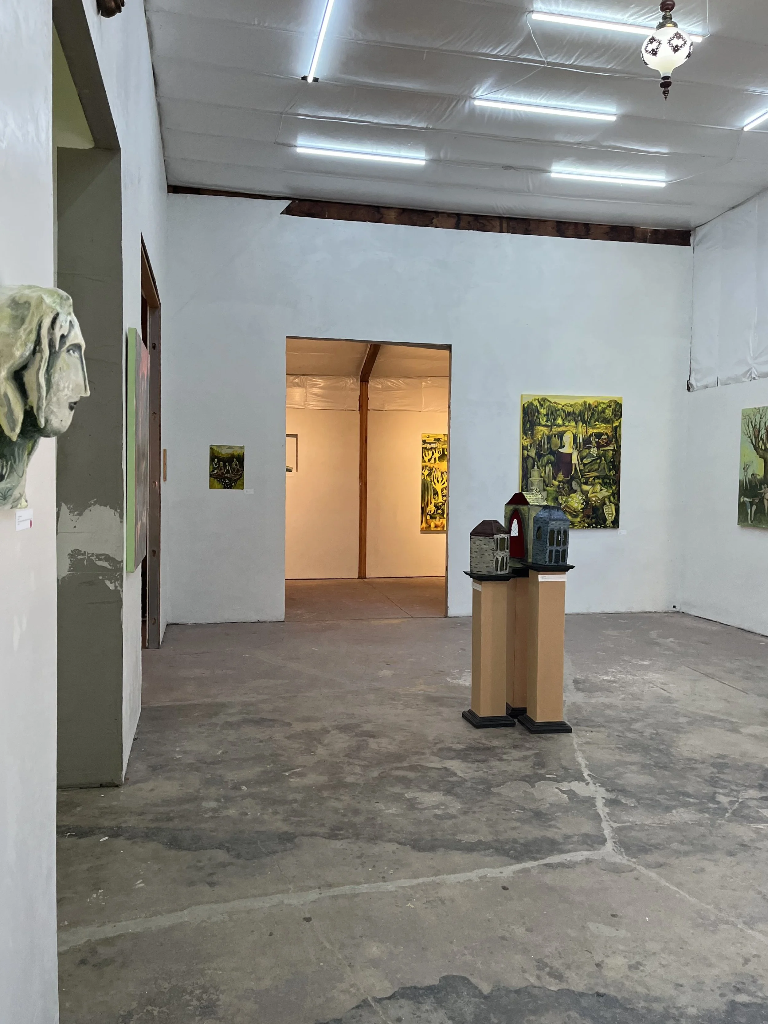 An art gallery inside a rustic building with white walls, artwork on display, and concrete floor. There is a pedestal with miniature models of houses and a wall-mounted sculpture of a face on the left.