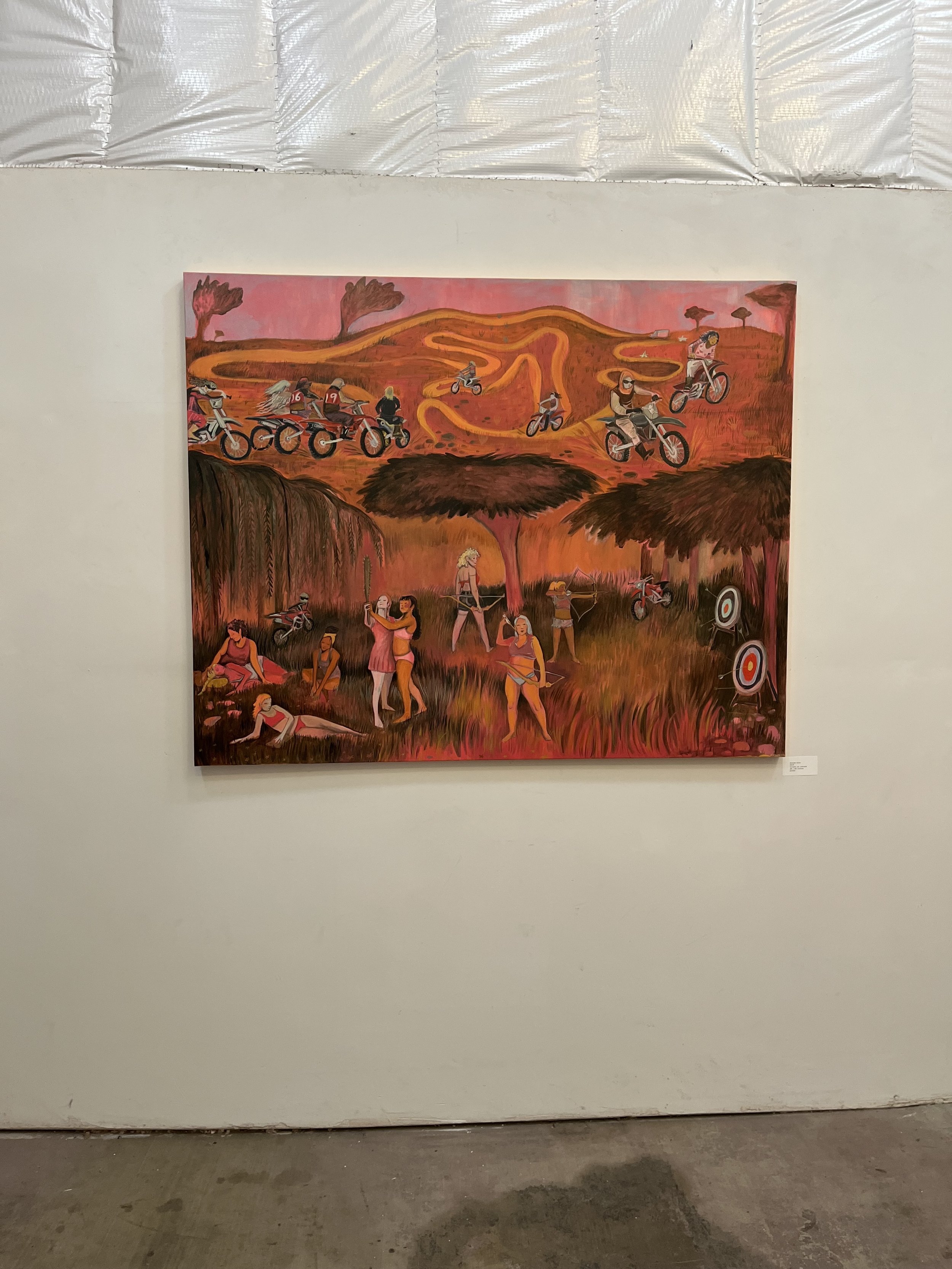 Painting on gallery wall depicting a group of people mountain biking on a orange and red landscape with trees and hills in the background.