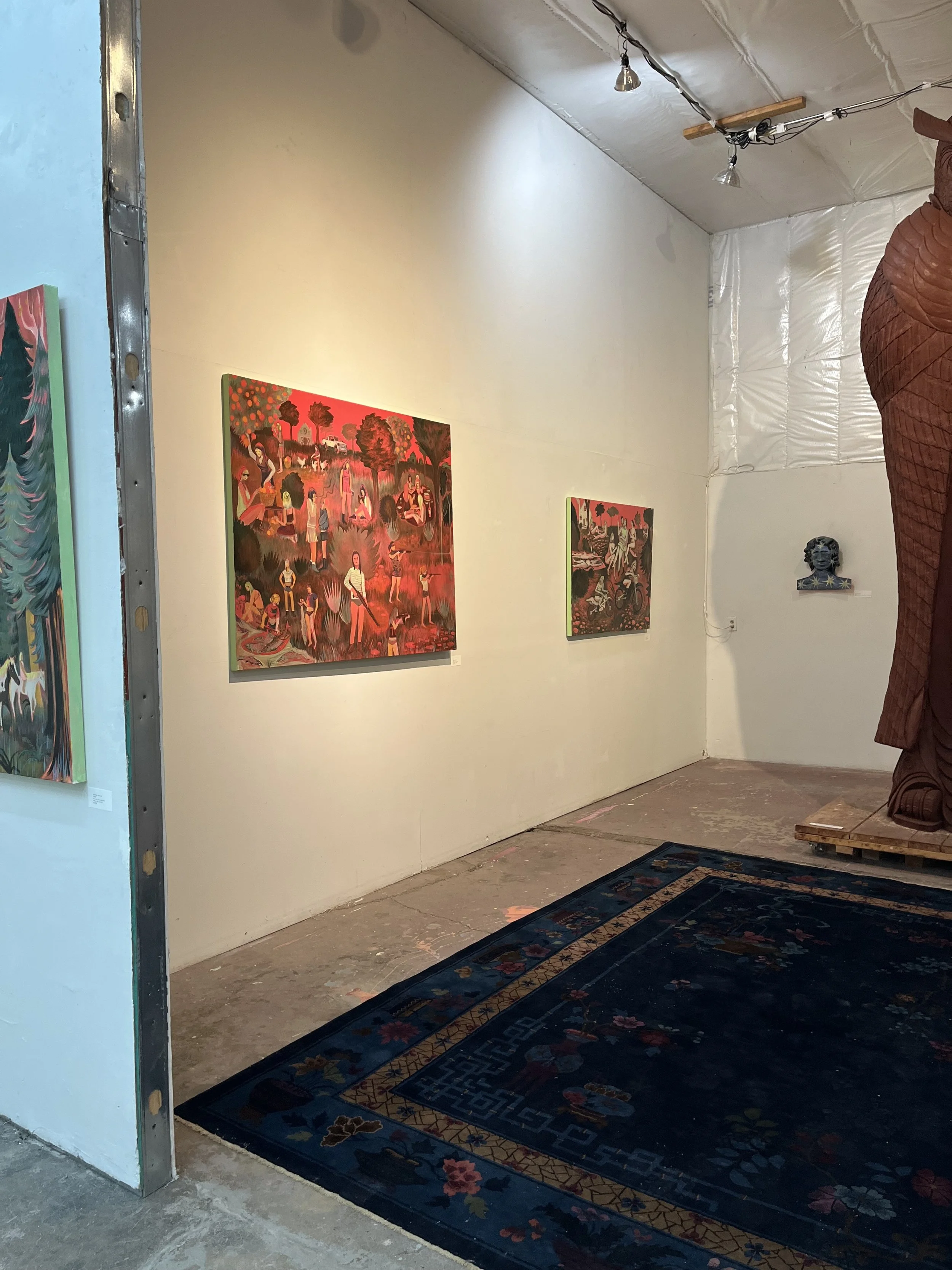 An art gallery wall displaying several colorful paintings with tree and human figures in an outdoor setting. Part of a patterned rug is visible on the floor in front of the wall.
