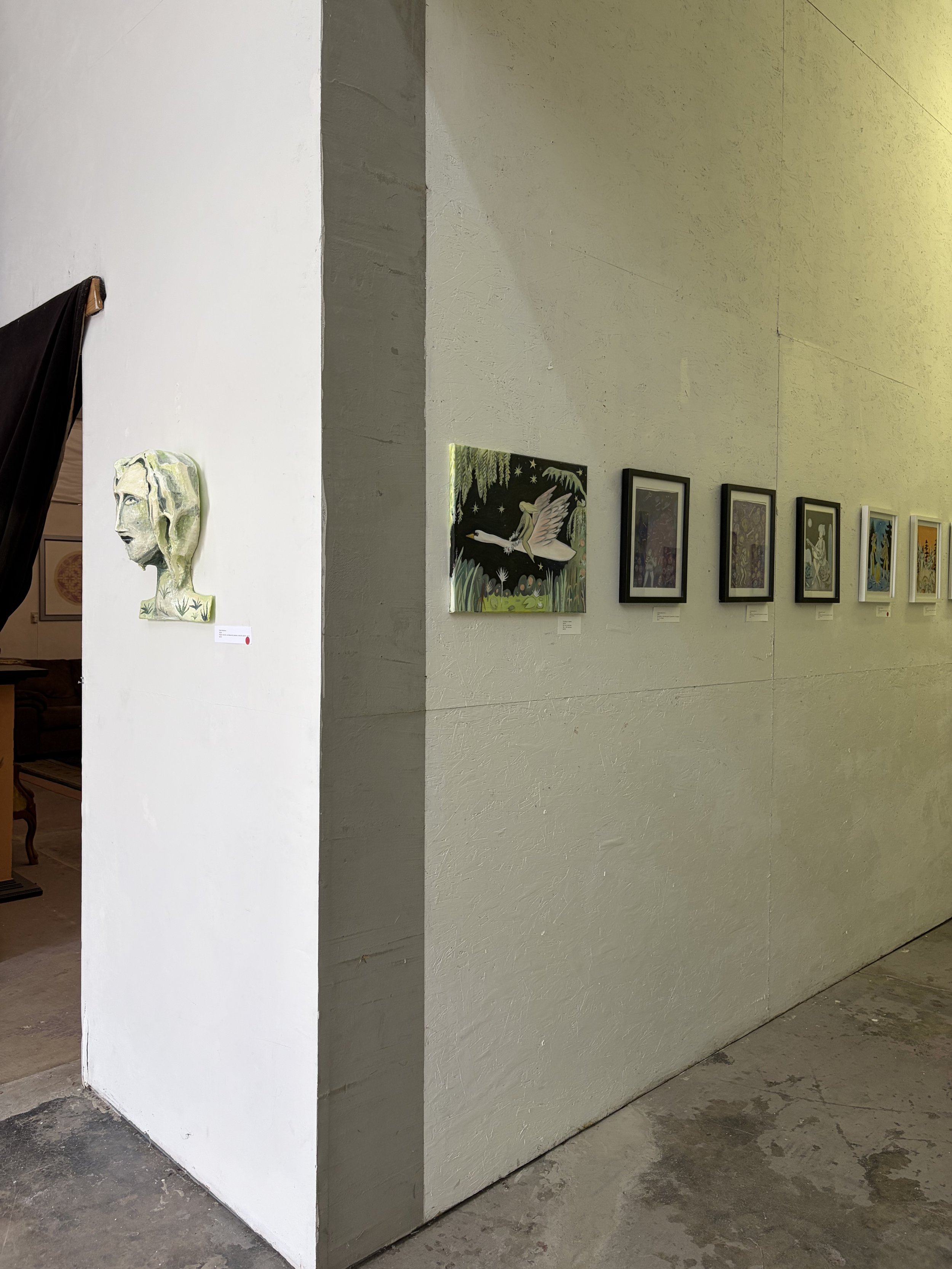 Art gallery wall featuring various paintings, including a portrait with long hair and a colorful artwork of a bird, with a concrete floor and a corner dividing the room.