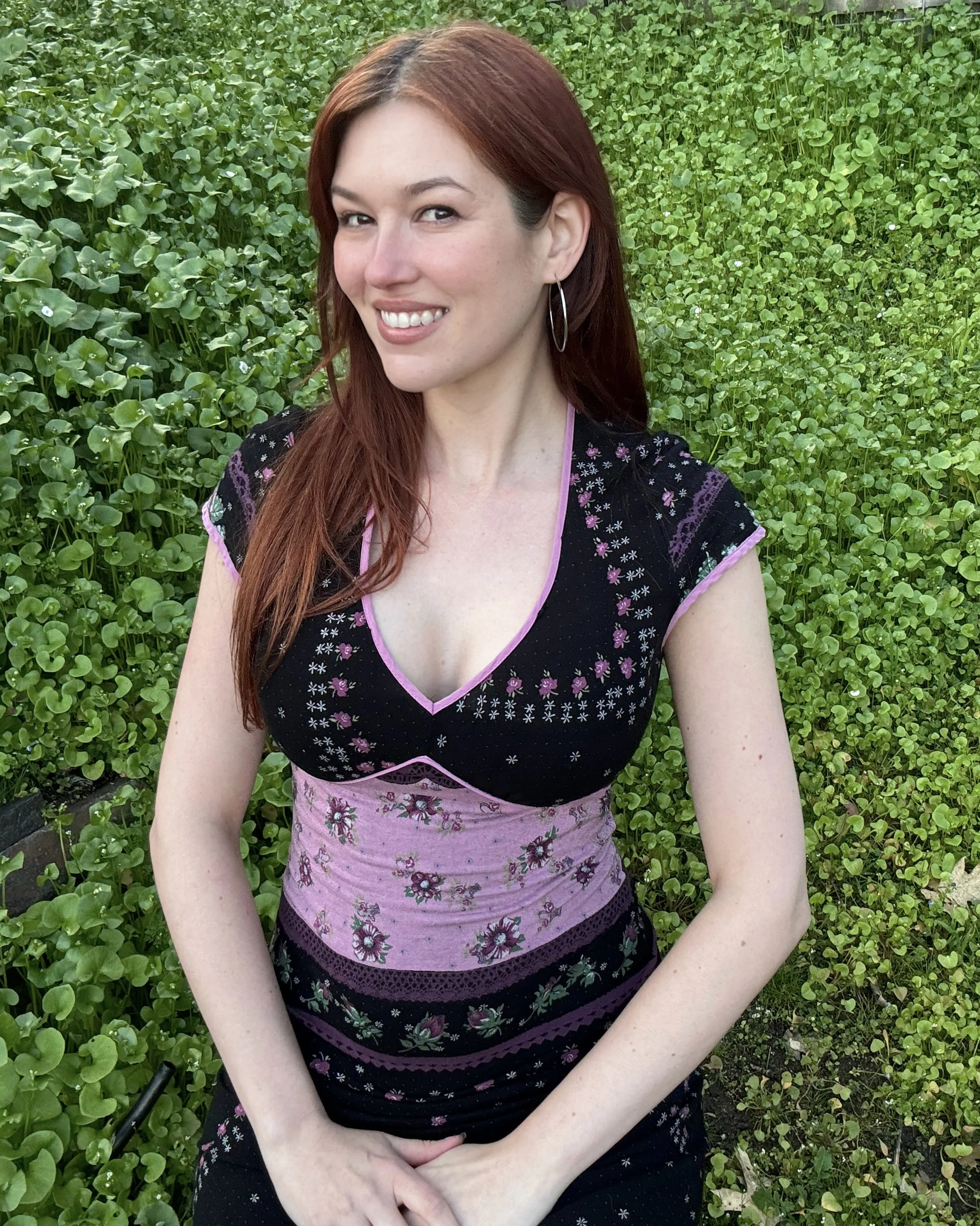Courtney Costa Artist, Courtney Costa Art, woman with long, reddish-brown hair smiling while sitting in a field of green clover. She is wearing a black top with floral embroidery and pink accents, and large hoop earrings.