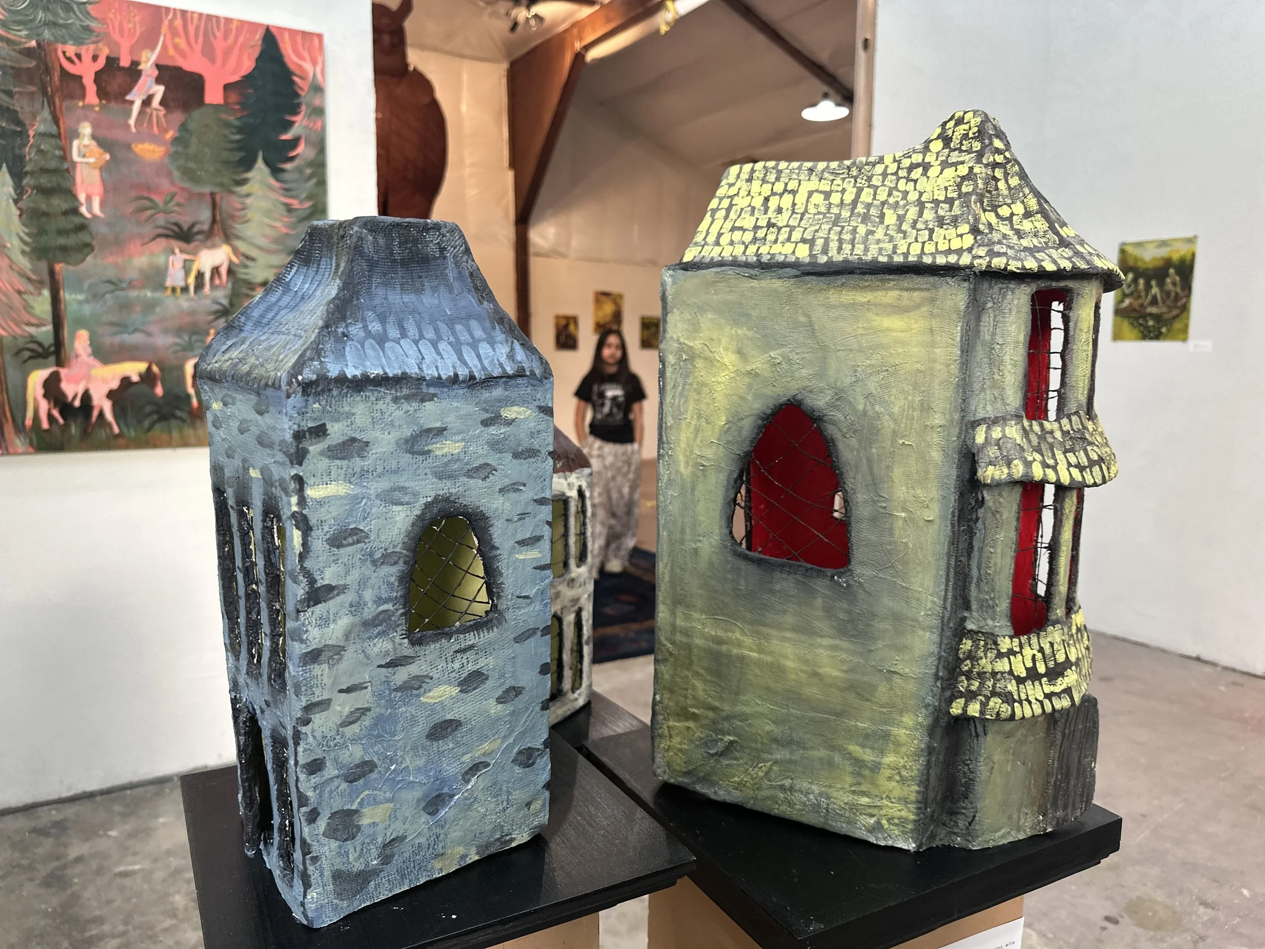 Two small, handcrafted ceramic houses with textured roofs, one blue and one yellowish-green, displayed on a black platform, with artwork and a woman visible in the background at an art gallery.