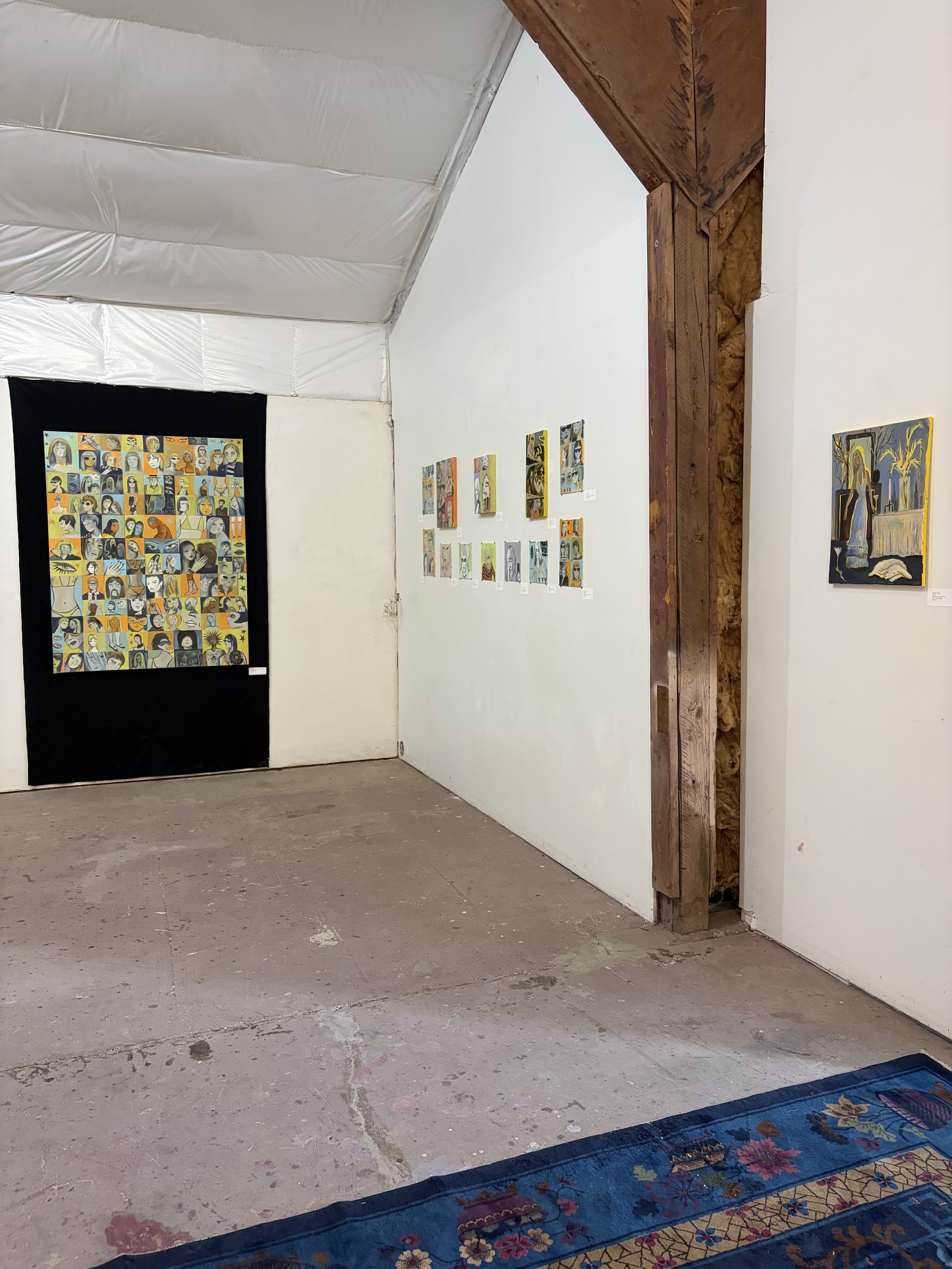 An art gallery with white walls displaying various paintings. The floor is concrete, and a colorful rug with floral patterns is partially visible at the bottom of the image. There is a large black frame on the left wall containing multiple small port