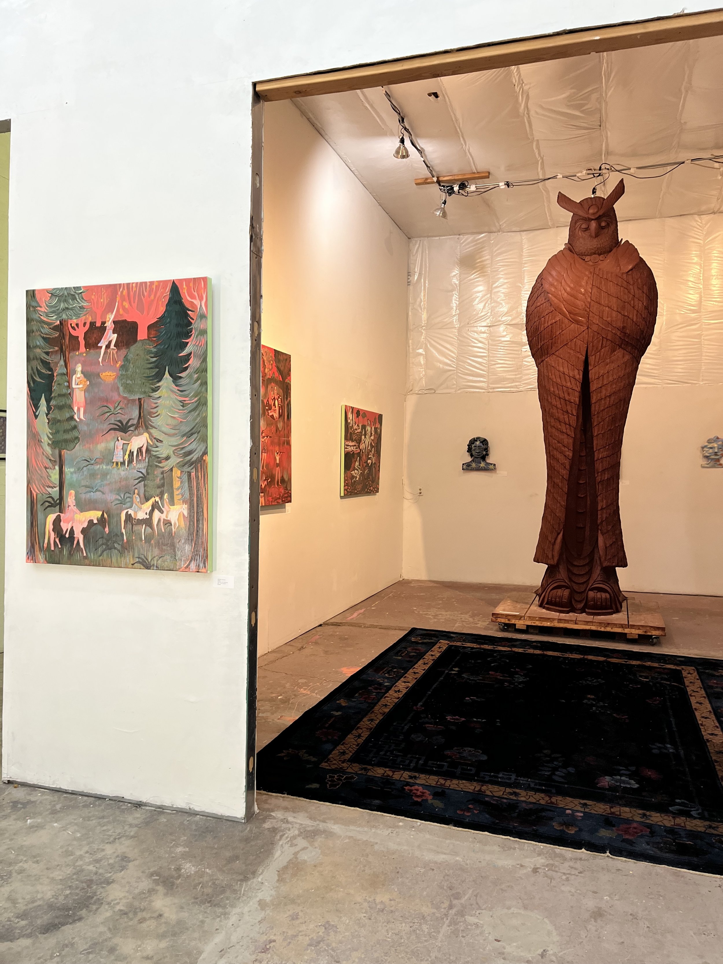 An art gallery exhibits a carved wooden owl sculpture on a wooden platform in a room with painted and artwork on the walls. The floor is concrete, and a black and red rug is in the foreground.