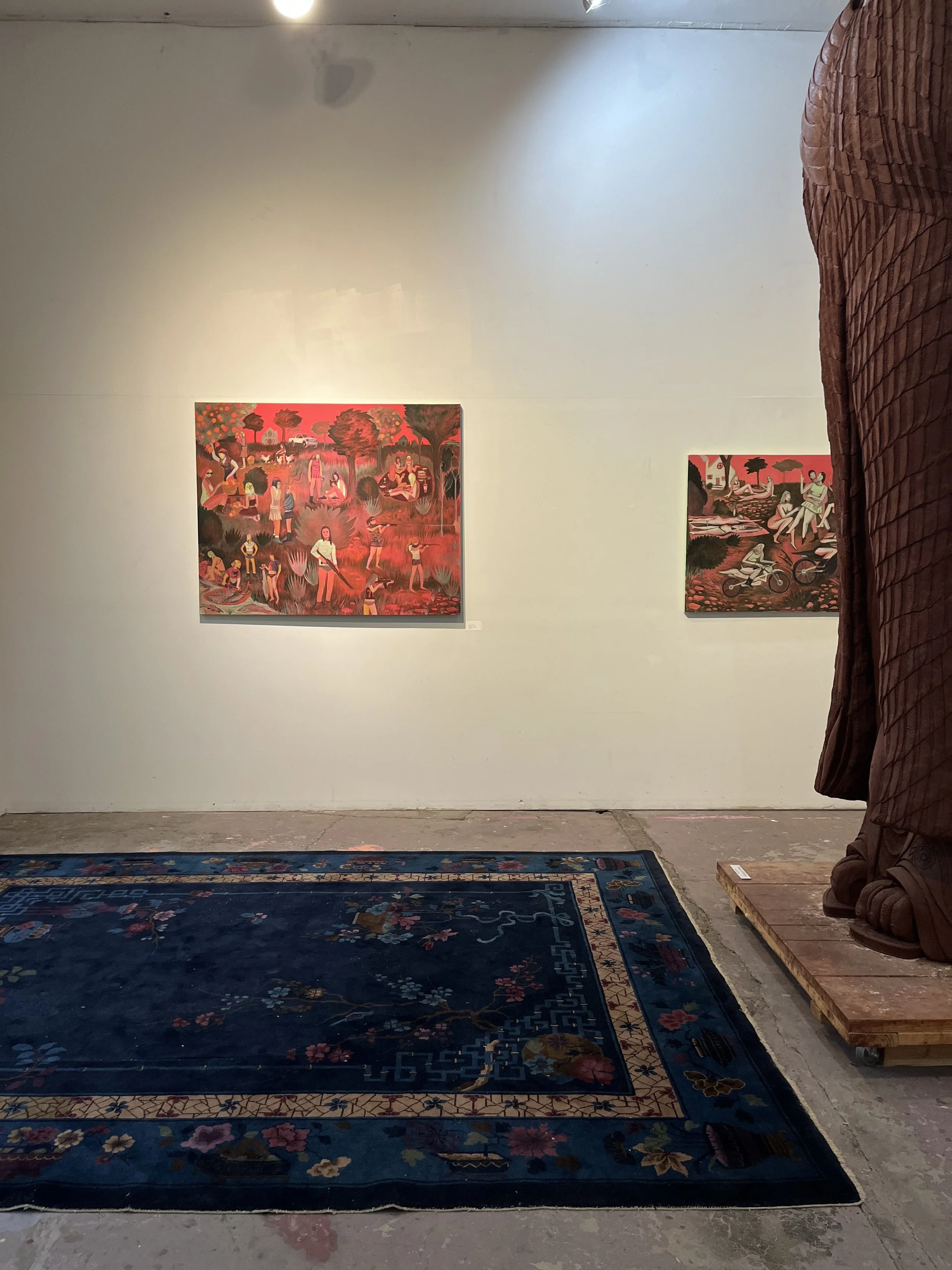 Gallery with two paintings on a white wall, a dark blue floral rug in the foreground, and a statue partially visible on the right side, draped in a brown fabric.