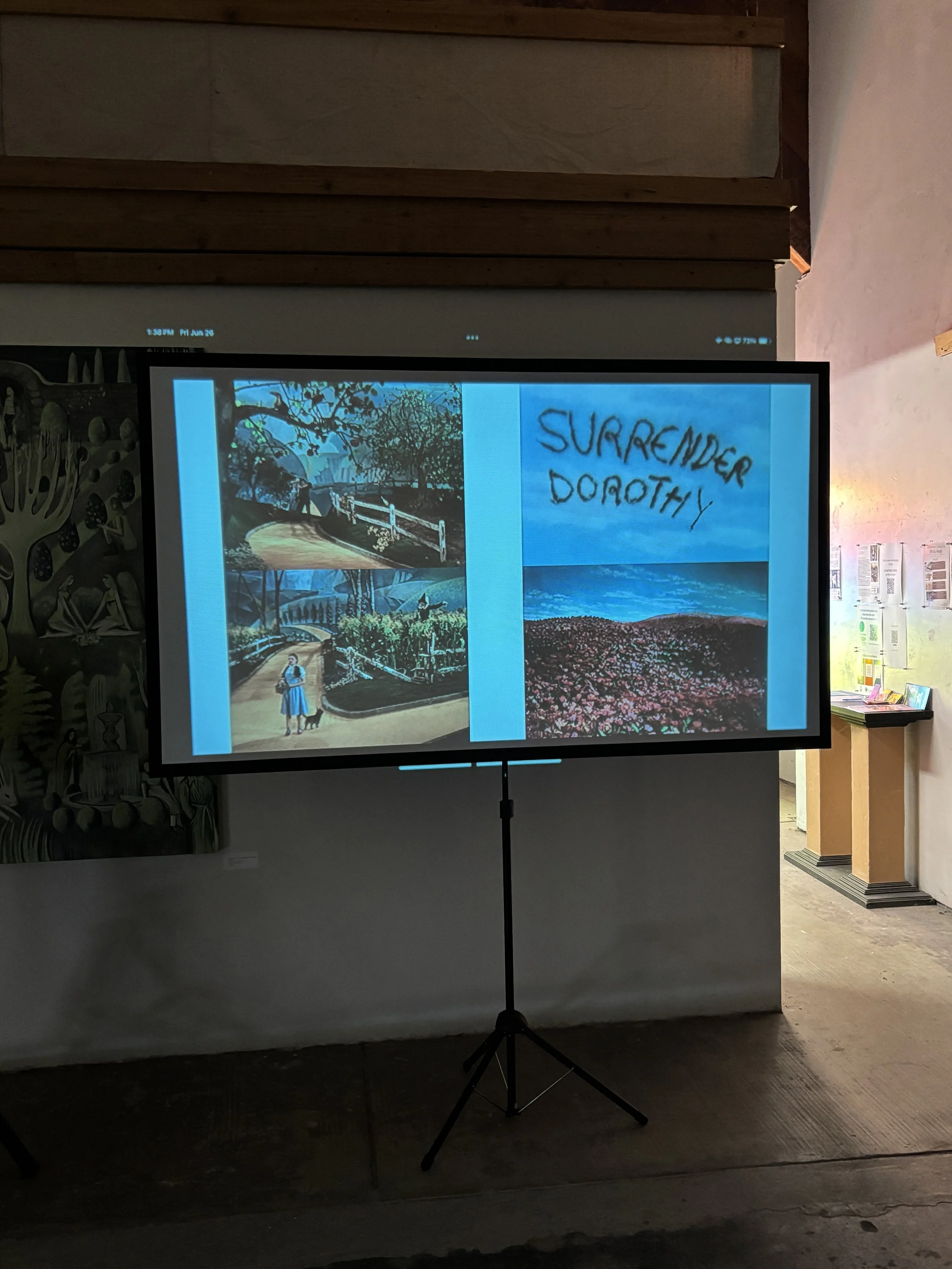 A digital screen displaying artwork and text in an art gallery with a dark floor, a white wall, and a wooden ceiling.