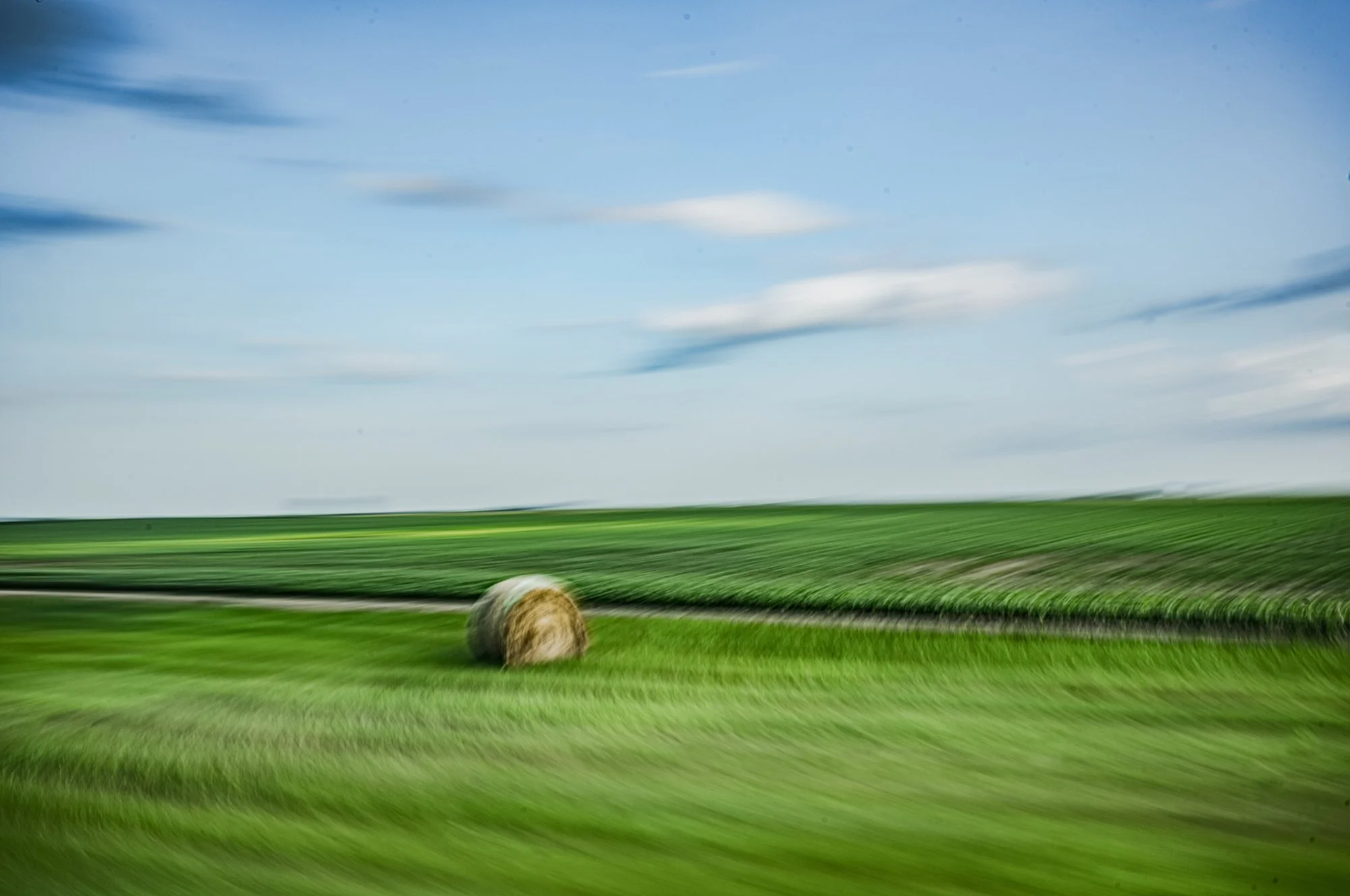 Photo of a hay bale in a green field under a partly cloudy blue sky.
