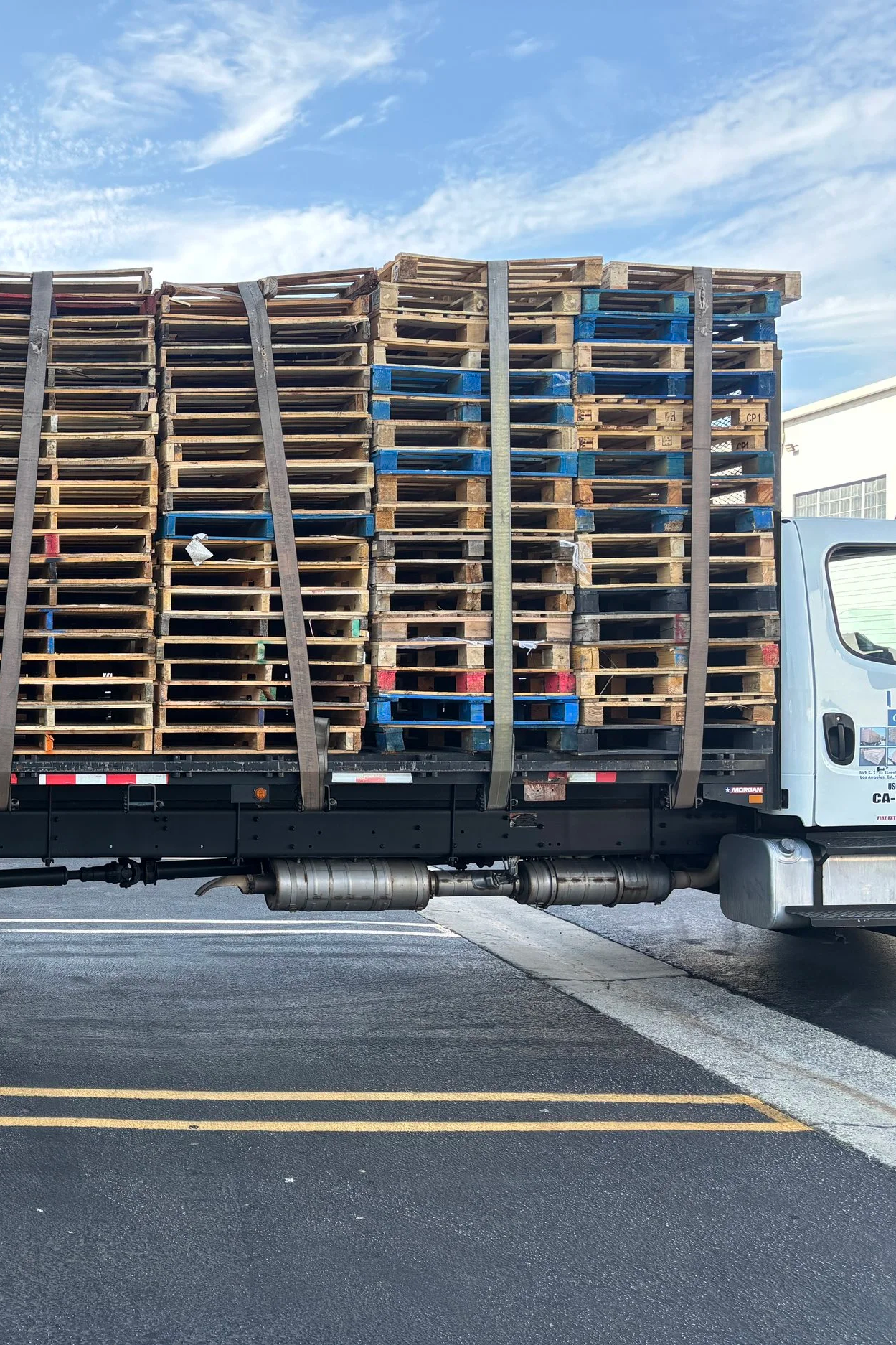 A truck loaded with stacked wooden pallets, secured with straps, parked on a paved lot against a partly cloudy sky.