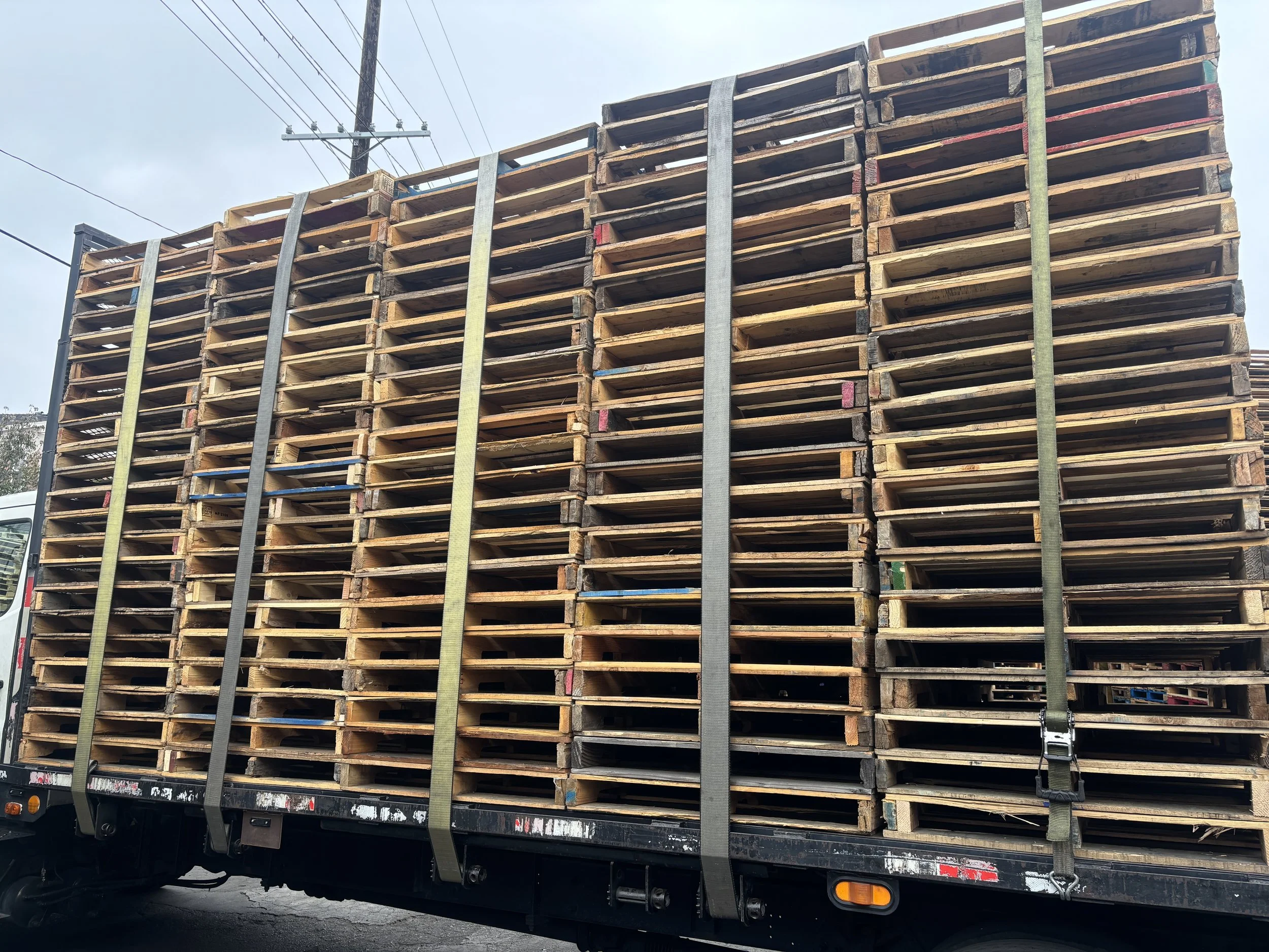 A load of wooden pallets stacked on a flatbed truck, secured with straps, with utility poles and wires in the background.