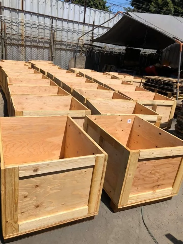 Multiple wooden plant or storage boxes outdoors, lined up in rows, with a fence and tent in the background.
