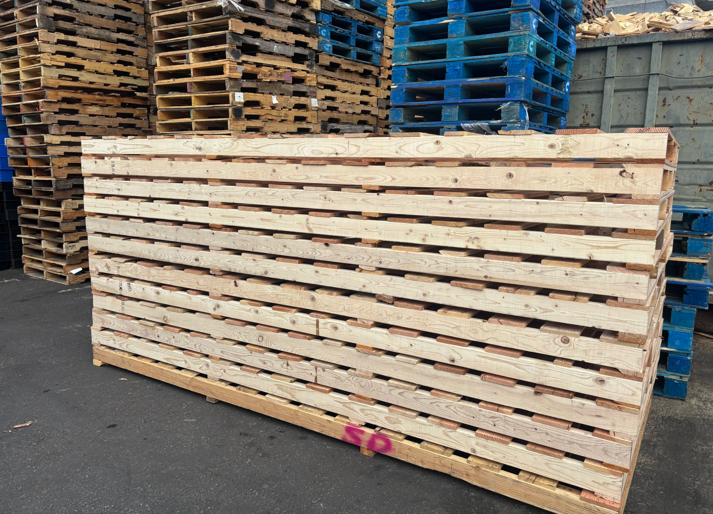Stacks of wooden pallets and crates stored outdoors on asphalt ground, with some pallets painted blue and others natural wood color.