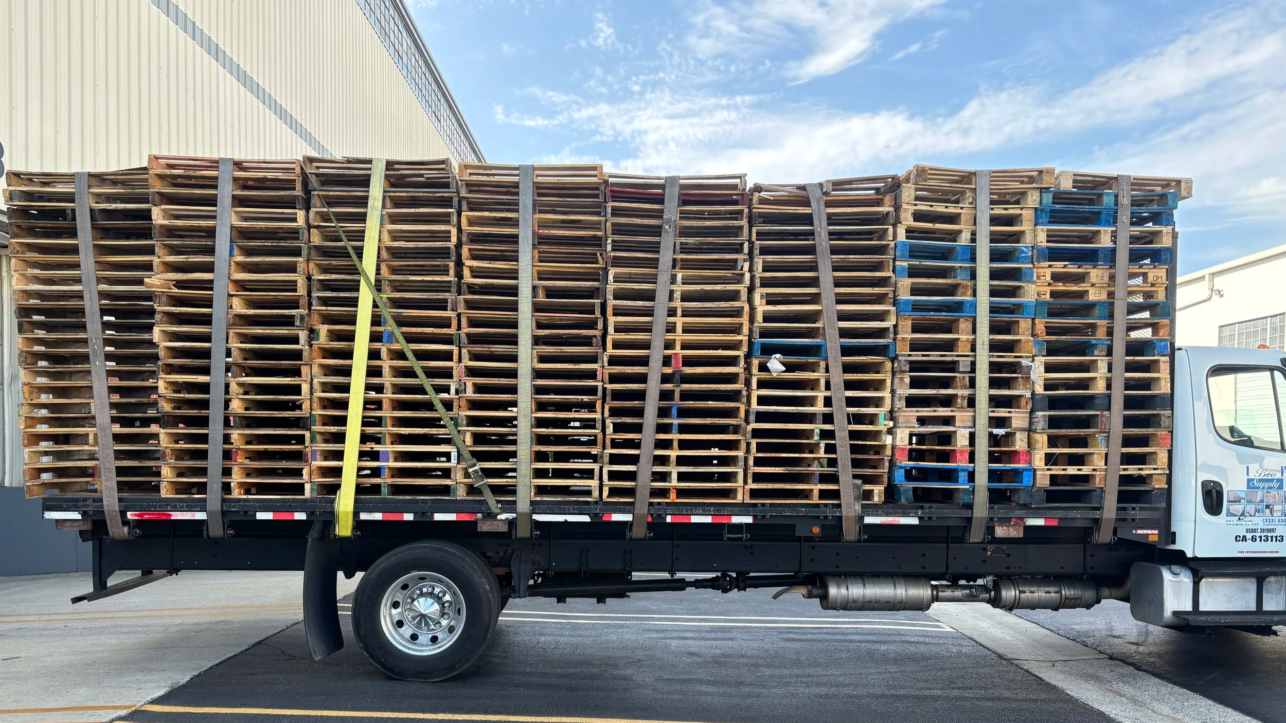 A flatbed truck carrying a large load of stacked wooden pallets secured with straps, parked in an industrial area with a building and blue sky in the background.