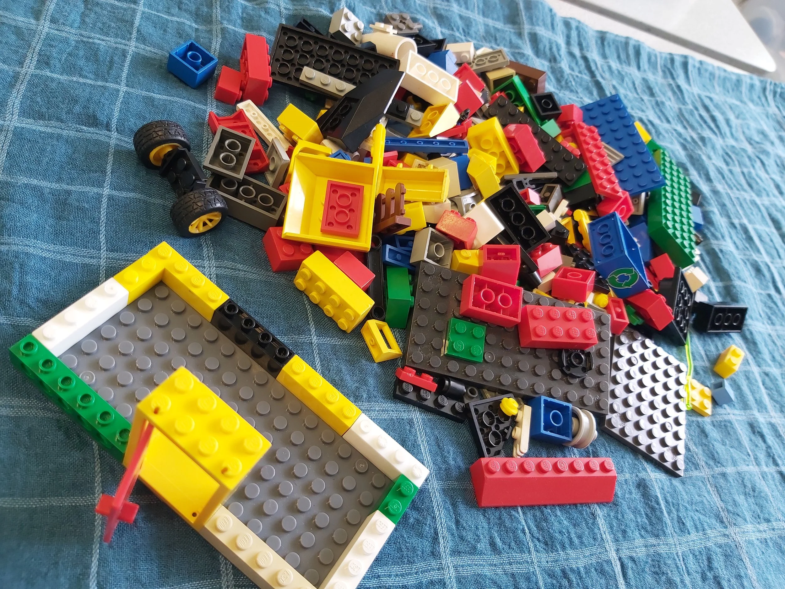 Pile of various colored LEGO bricks scattered on a blue checkered fabric surface, including wheels, plates, and blocks in red, yellow, black, green, white, and gray.