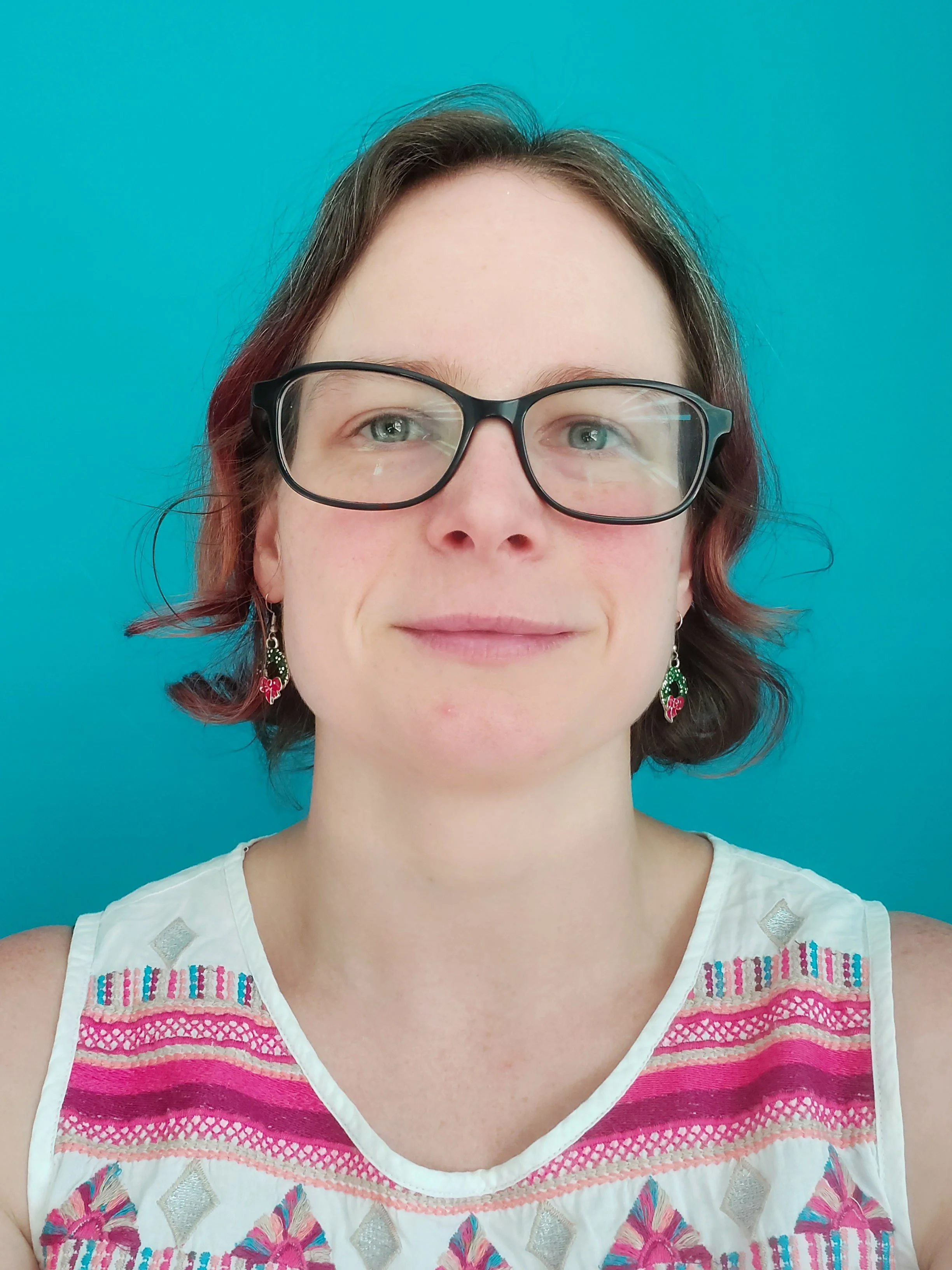 A woman with short brown hair, wearing black glasses, earrings, and a white embroidered top with pink, blue, and silver designs, standing against a bright blue background.