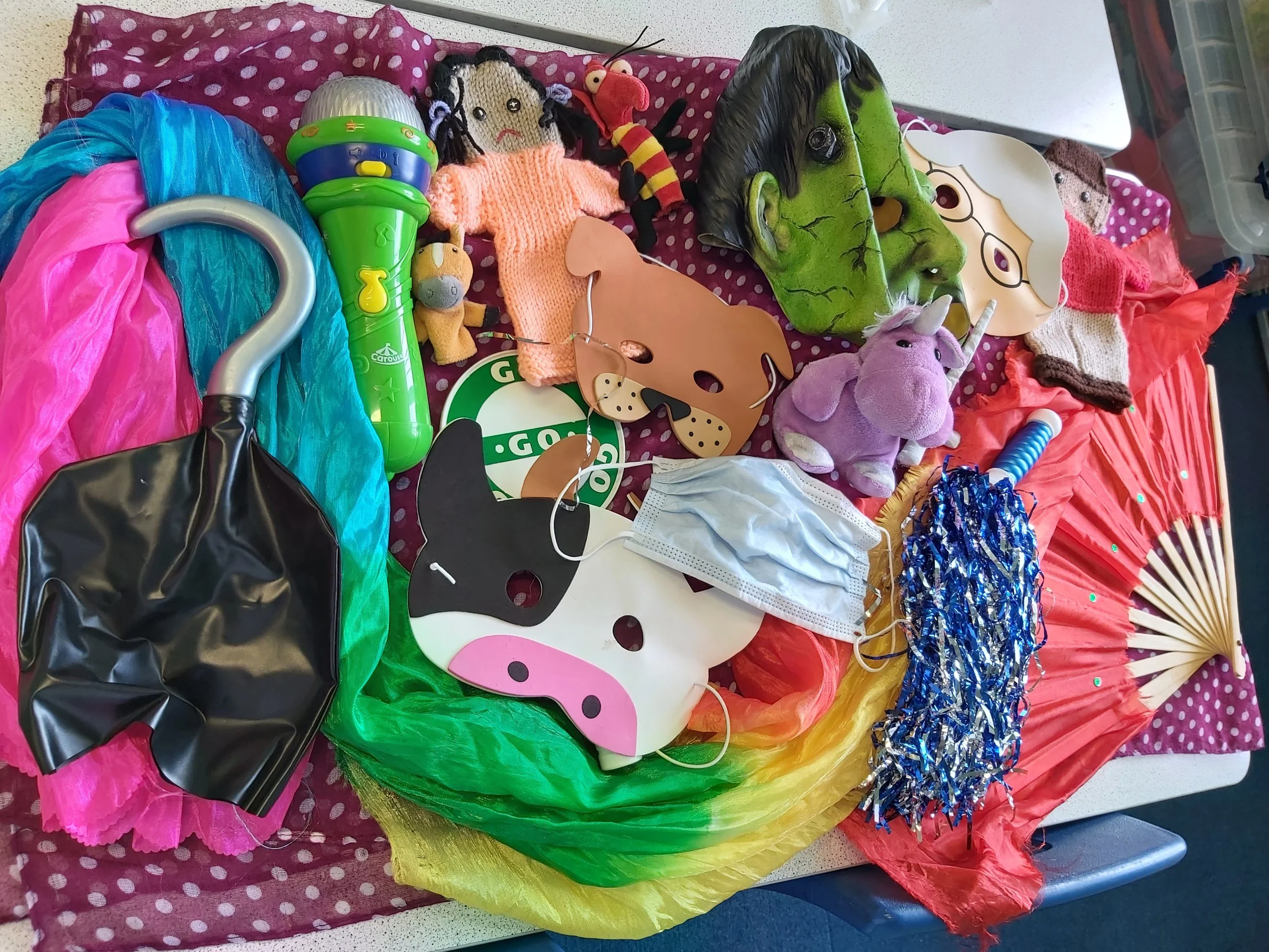 Colorful assortment of toys, costumes, and decorations on a polka-dotted cloth, including a witch mask, a unicorn plush, a toy guitar, a doll, a maraca, and various pieces of fabric and accessories.