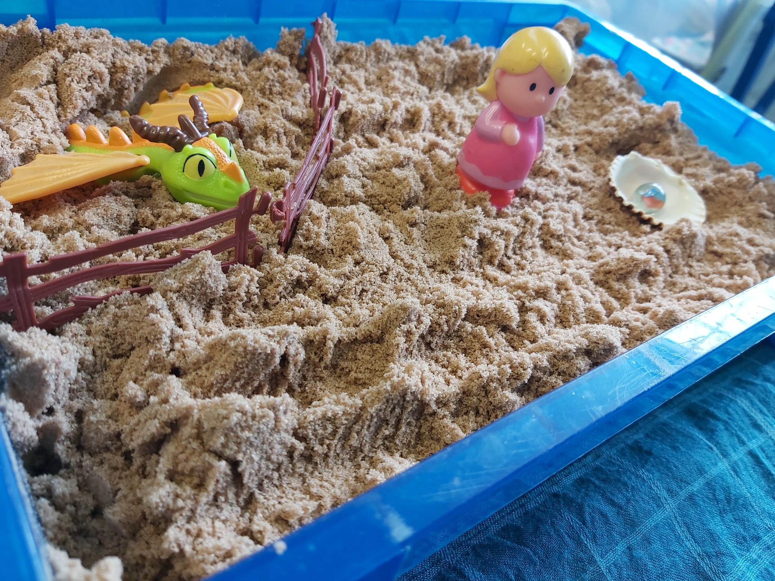 Toy dinosaur and a toy girl figure in a sandbox with sand and a small plastic fence.