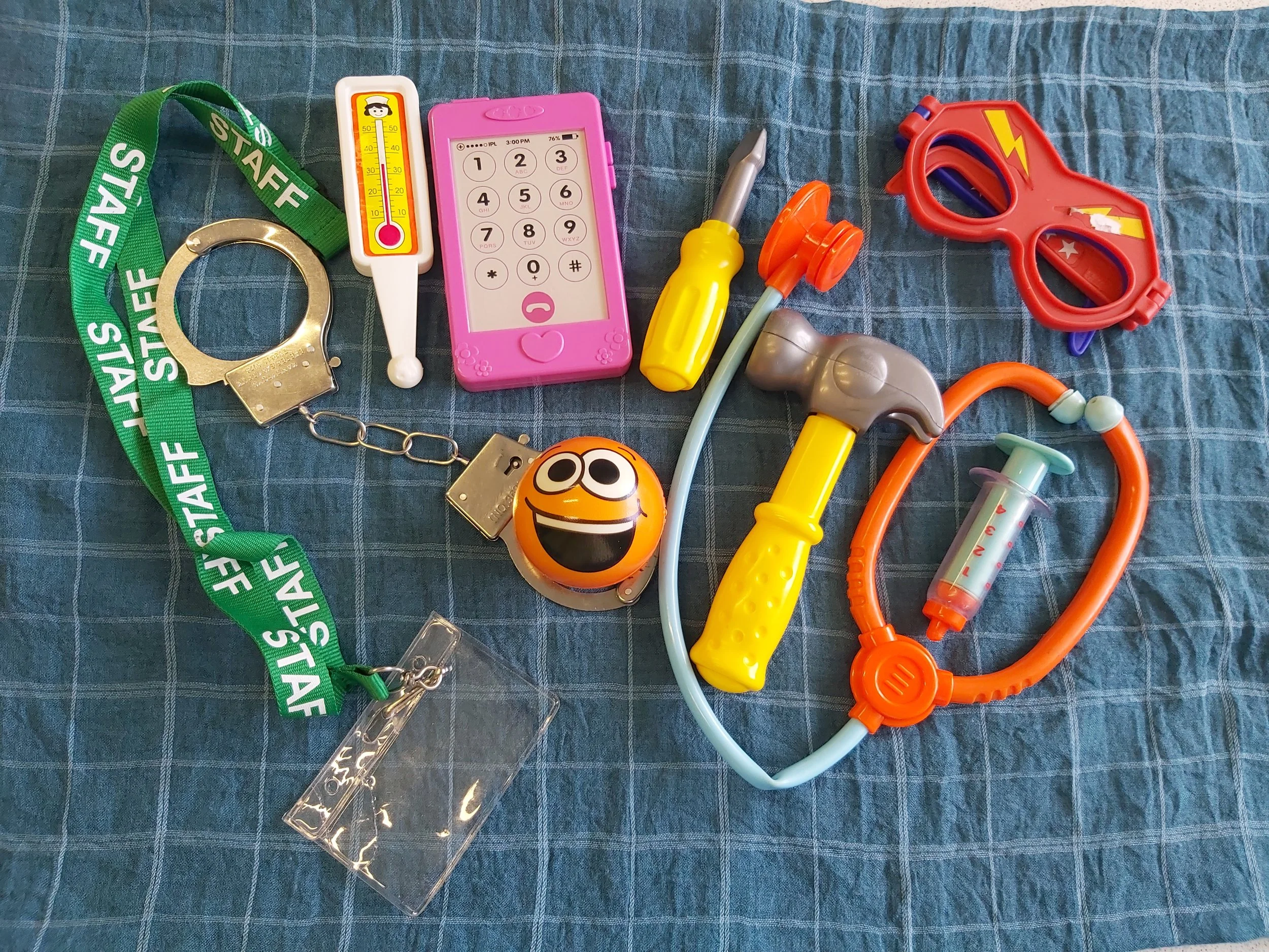 Collection of toy and novelty items on a blue checkered fabric surface, including a green 'STAFF' lanyard, a metal handcuff, a pink toy phone, a yellow toy screwdriver, a red toy laser gun, a laughter emoji ball, a plastic brain model, and various colorful plastic tools.