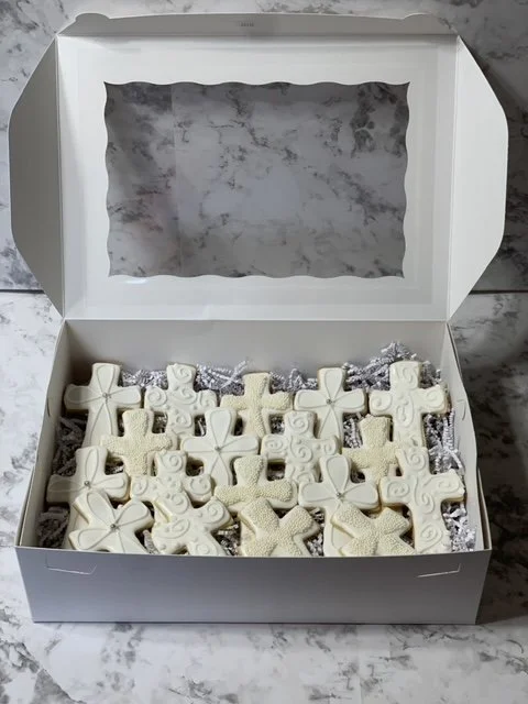 A white box containing a variety of white and cream decorative soap bars shaped like crosses and stars, wrapped in silver and purple tissue paper.