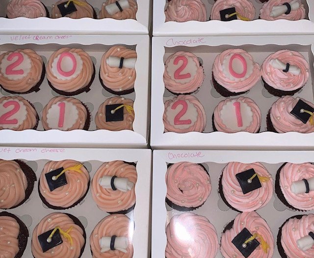 Six boxes of cupcakes decorated for New Year's celebration with pink and chocolate frosting, themed for 2021, featuring cake toppers of the numbers 2, 0, 2, 1, and cupcakes with graduation cap toppers and scroll decorations.