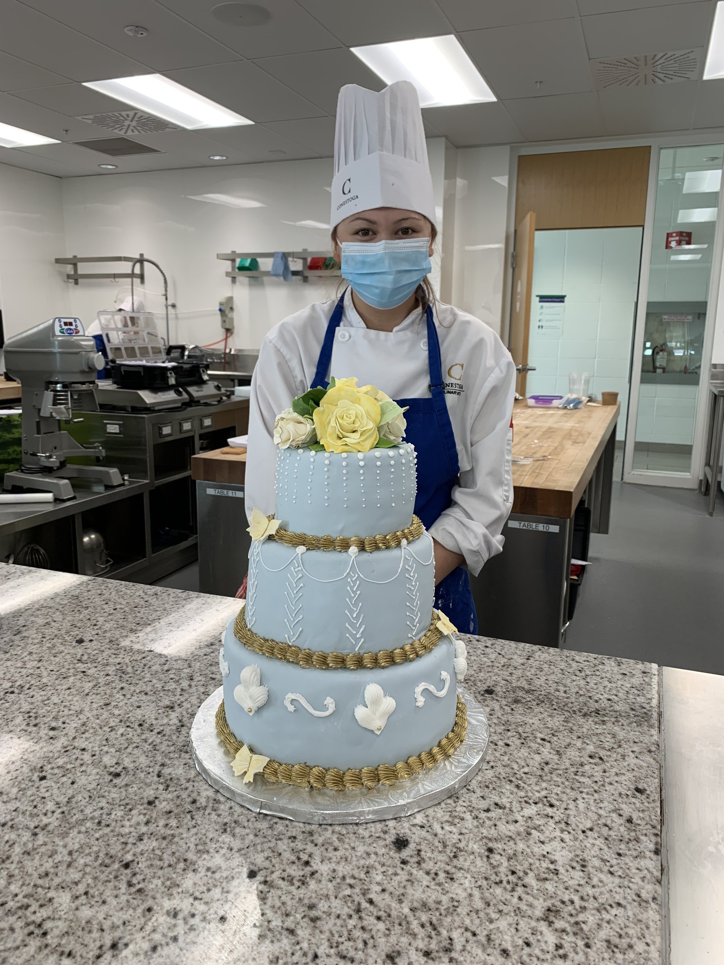 A woman dressed as a chef, wearing a tall chef hat and a face mask, proudly displays a large, multi-tiered white cake decorated with flowers and gold accents in a commercial kitchen.