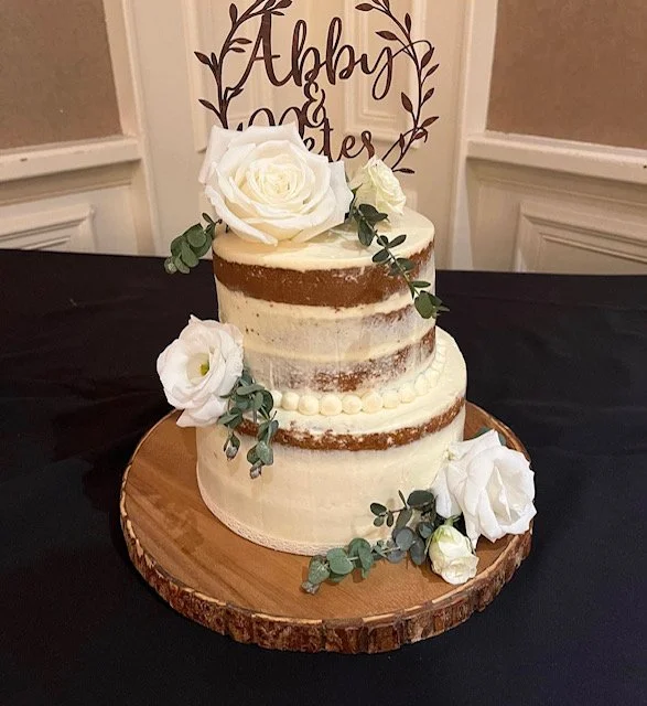 A two-tier semi-naked wedding cake decorated with white roses and green foliage, placed on a wooden cake board, with a sign in the background reading 'Abby and Peter'.