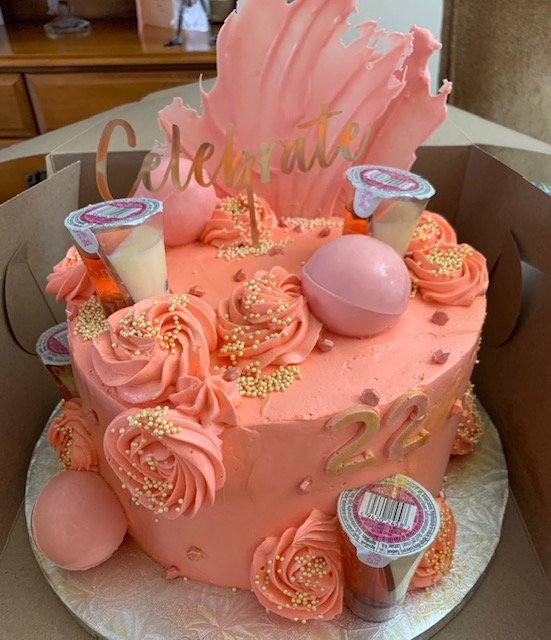 A pink birthday cake decorated with pink frosting rosettes, sprinkles, edible pearls, and small drink-shaped toppers. It also features a gold 'Celebrate' topper, a pink decorative block, and the number '22'. The cake is in a brown box.