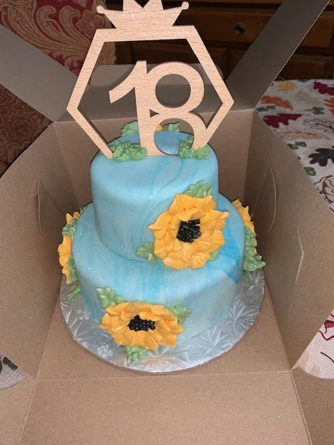 Two-tiered cake with blue icing, decorated with yellow sunflowers and green leaves, inside a cardboard box. Cake topper features a wooden cutout with a crown and a logo with the number 10.