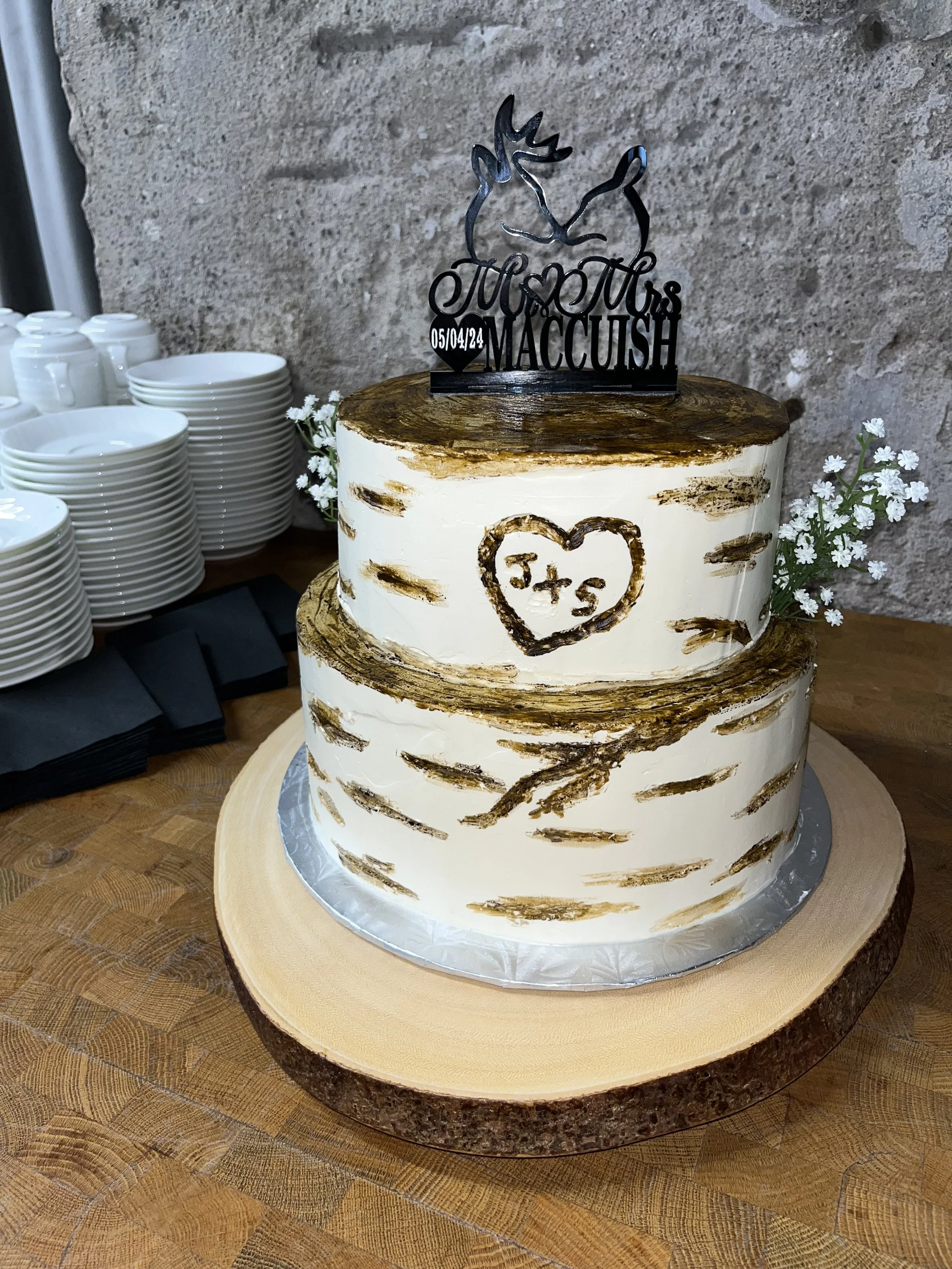 A two-tiered wedding cake with a rustic birch bark design, topped with a deer and antlers cake topper that says 'Just Married' and includes a date, placed on a wooden slab, with white flowers and stacks of white plates nearby.
