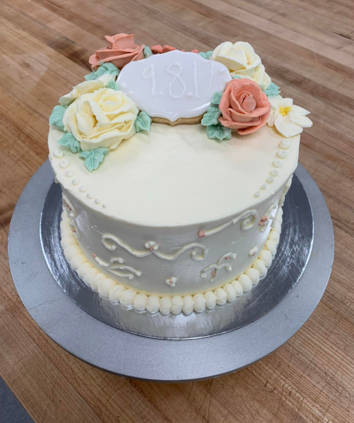 Round white anniversary cake decorated with pastel-colored flowers and a plaque with the date 18.1, on a silver cake board.