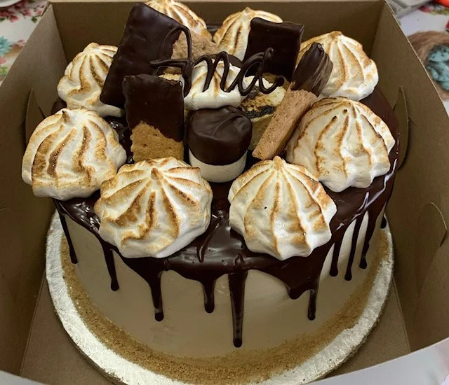 A round cake with chocolate drizzle and toasted marshmallows on top, decorated with assorted chocolates, cookies, and a chocolate "No" sign.