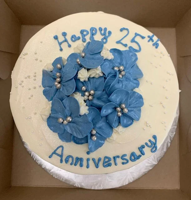 Round white cake with blue and white flower decorations, silver beads, and blue writing that says 'Happy 25th Anniversary'.