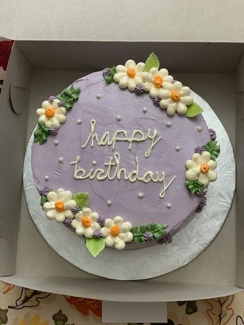 Round birthday cake with purple icing, decorated with white and orange flowers and green leaves, with 'happy birthday' written in white icing in the center.