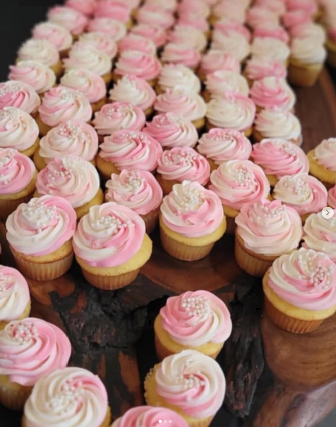 Numerous cupcakes with pink and white swirled frosting topped with pearl-like sprinkles on a wooden surface.