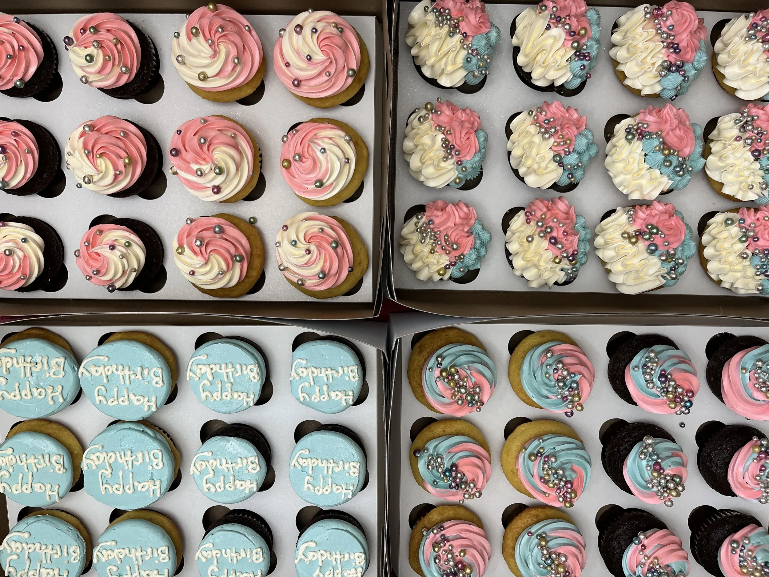 A display of cupcakes in boxes with pink, white, and blue frosting decorated with multicolored edible beads and sprinkles, some with birthday messages.
