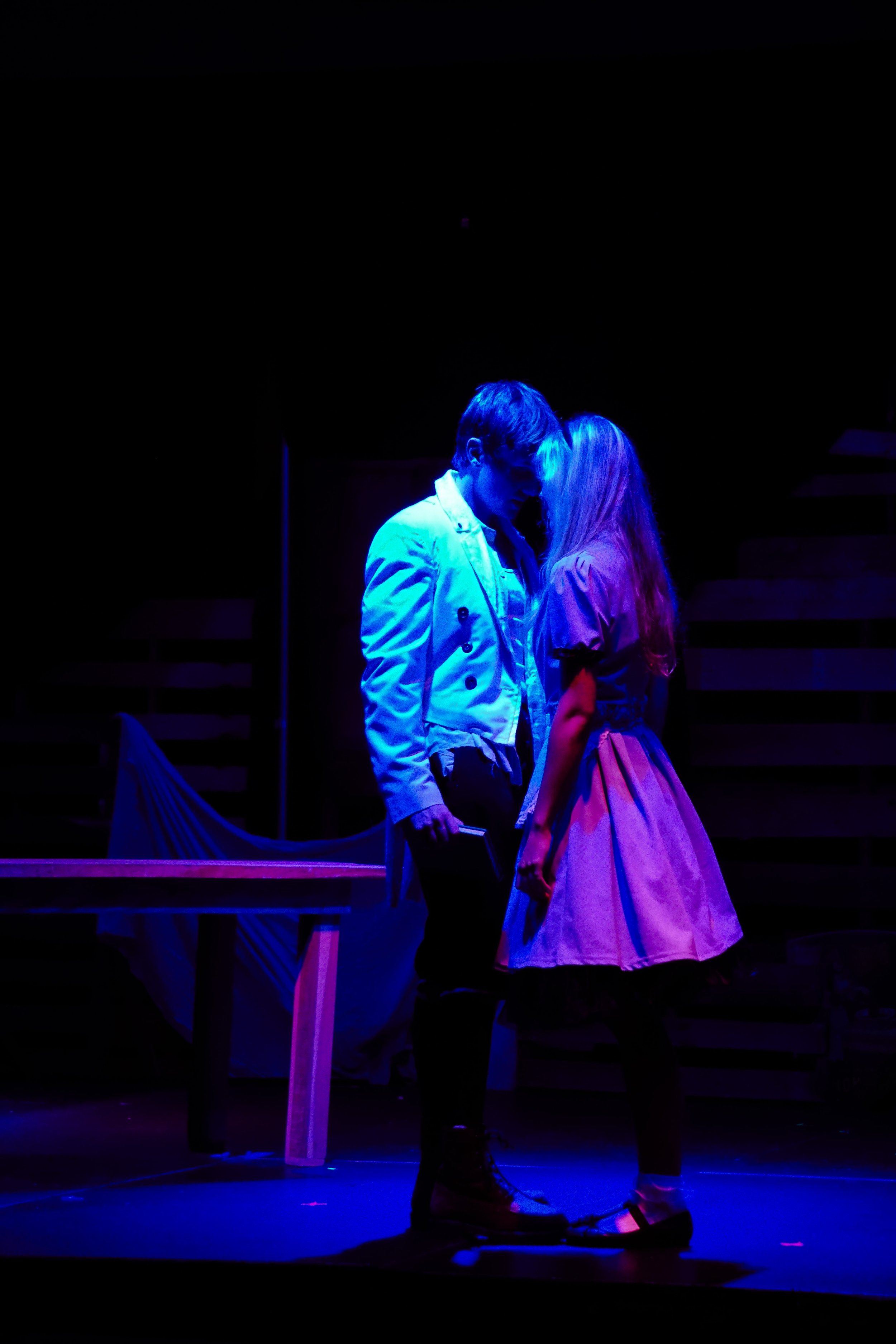 Two performers, a man and a woman, stand closely together on a dark stage with purple and blue lighting. The man wears a light-colored jacket, and the woman wears a light-colored dress. They are facing each other with their foreheads touching, creating an intimate moment.