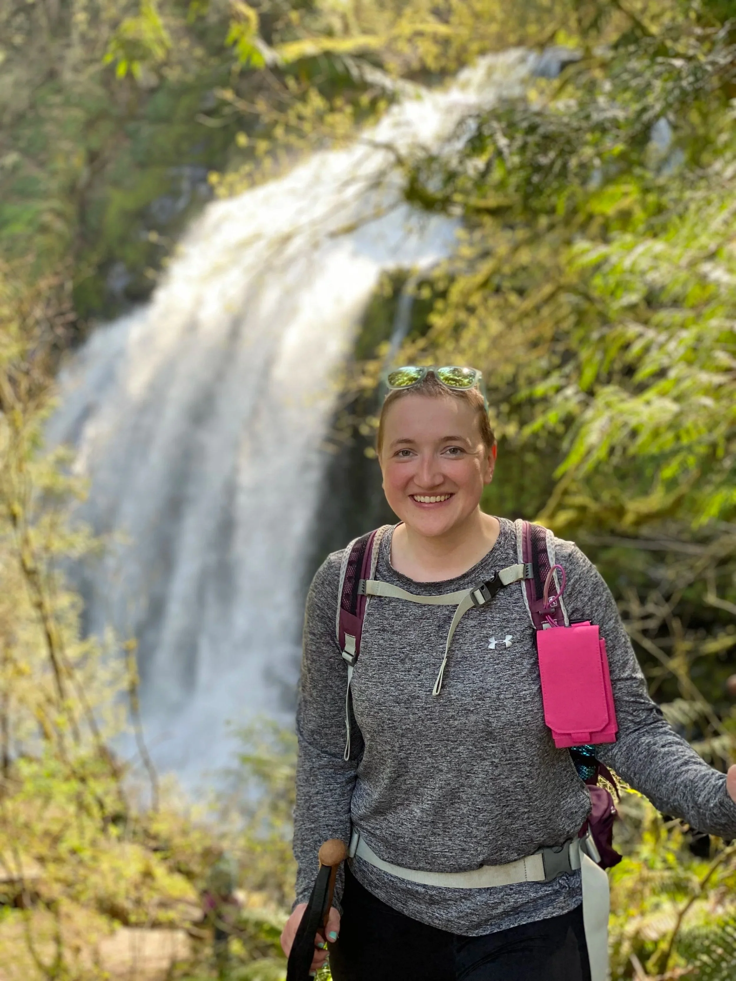 A woman smiling outdoors in front of a waterfall, wearing sunglasses on her head, a gray long-sleeve shirt, and a backpack, holding a hiking pole.