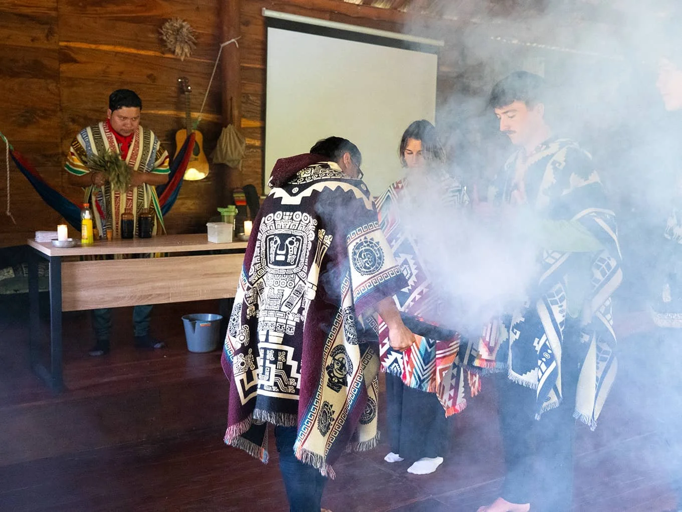 People dressed in traditional indigenous clothing perform a ceremonial ritual indoors, with smoke and a colorful woven textile, while others observe near a wooden wall with candles and decorative items.