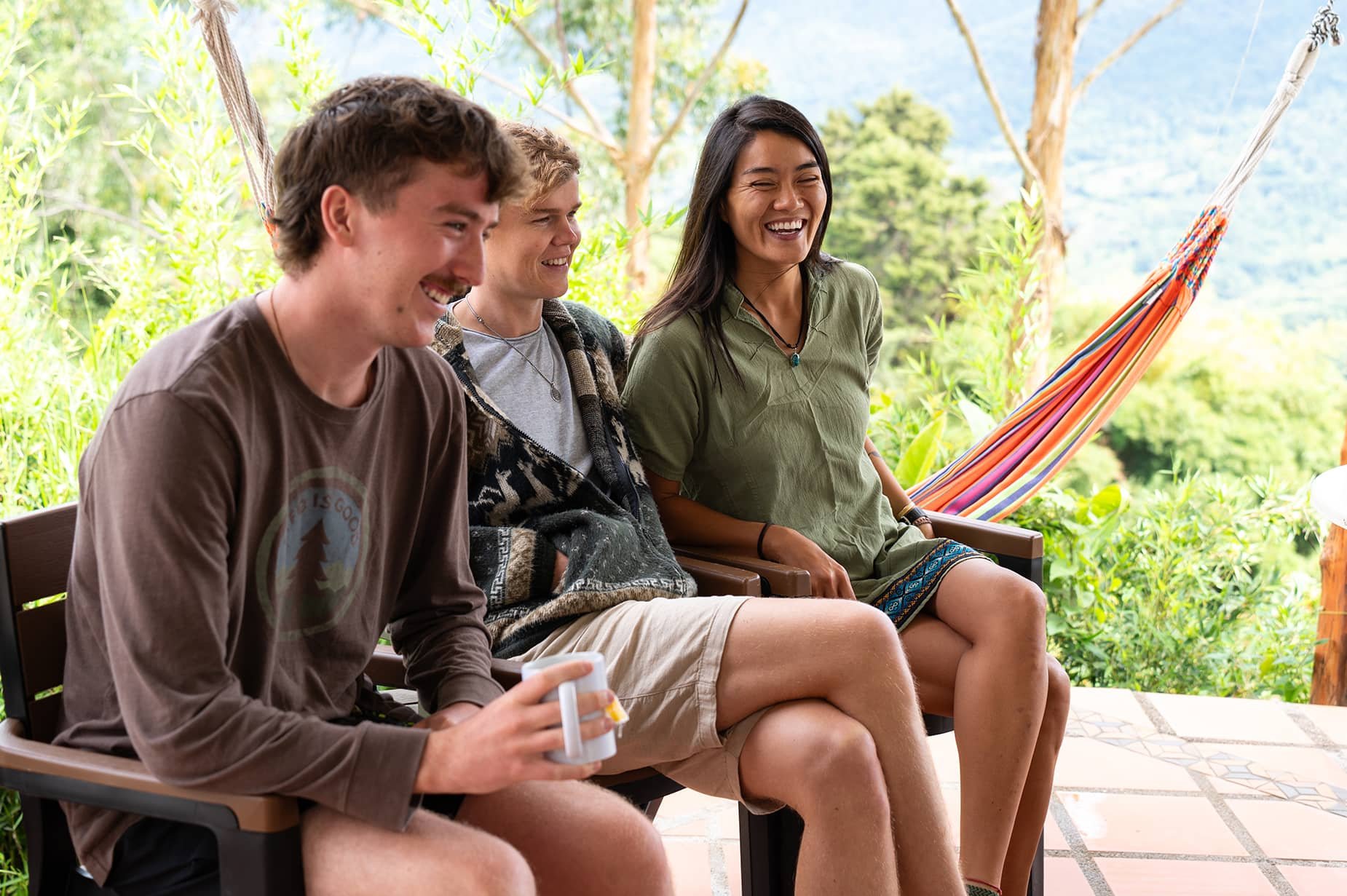 Four young adults sitting on a porch, smiling and laughing. One person is holding a white mug, with a colorful hammock and green foliage in the background.