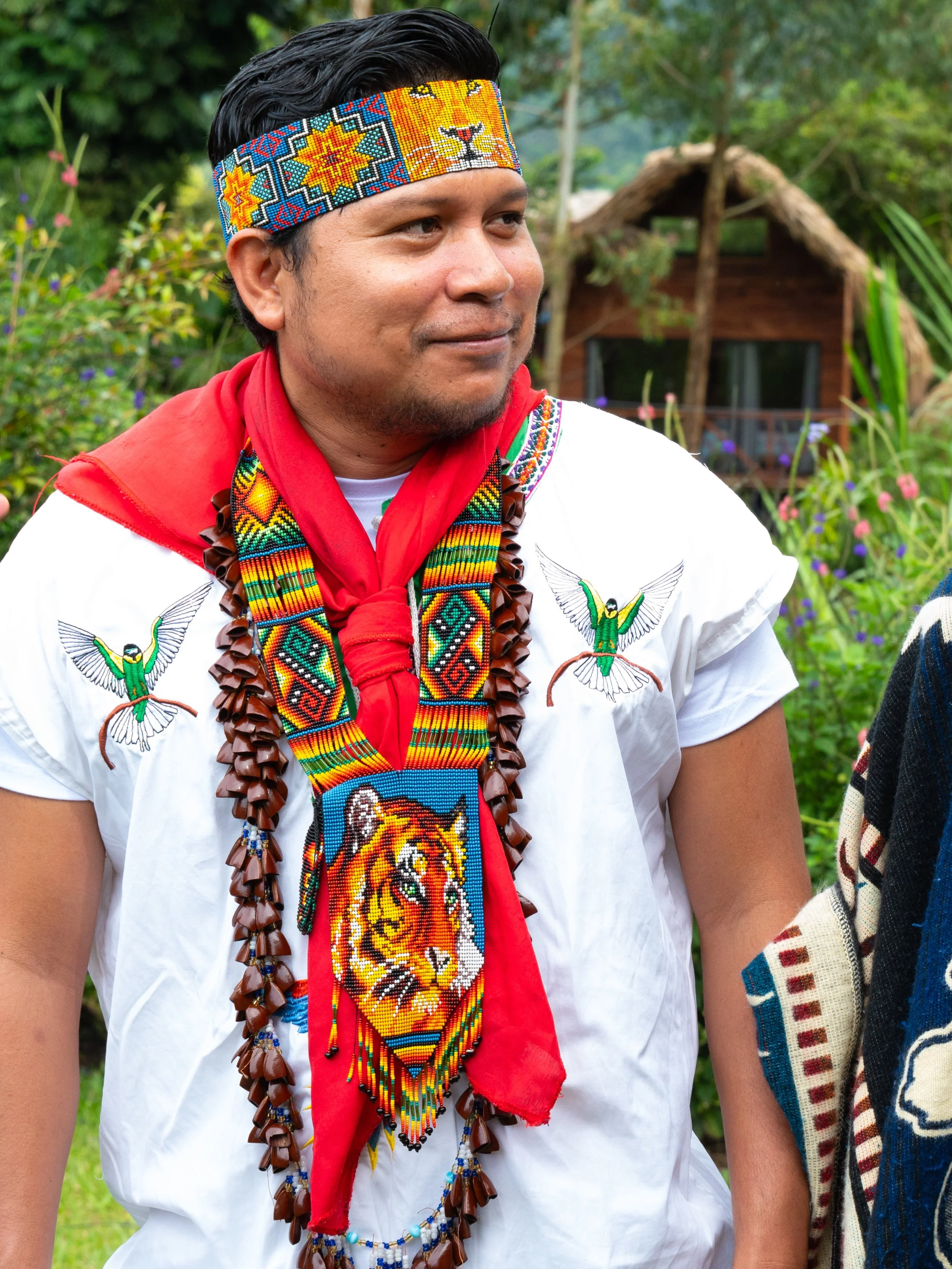 A man wearing traditional indigenous clothing with beaded jewelry and a beaded headband, standing outdoors among greenery.