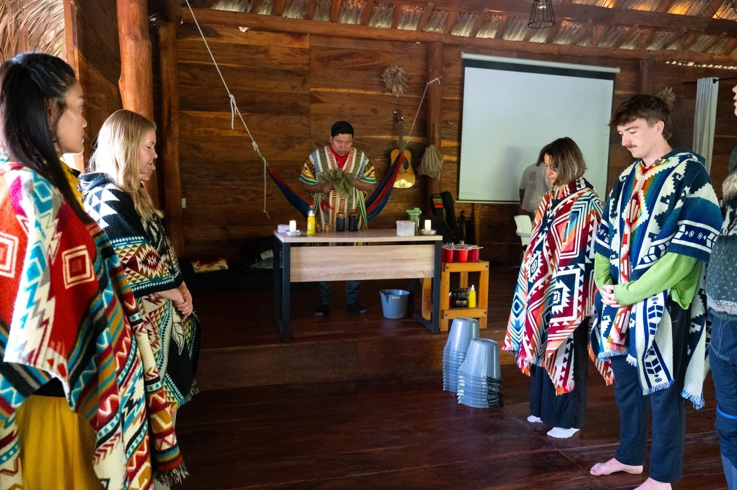 Group of people standing in a circle inside a wooden room, wrapped in colorful tribal blankets, participating in a ceremonial or spiritual ritual with a person in front of them holding herbs, in a setting that resembles a sauna or native lodge.