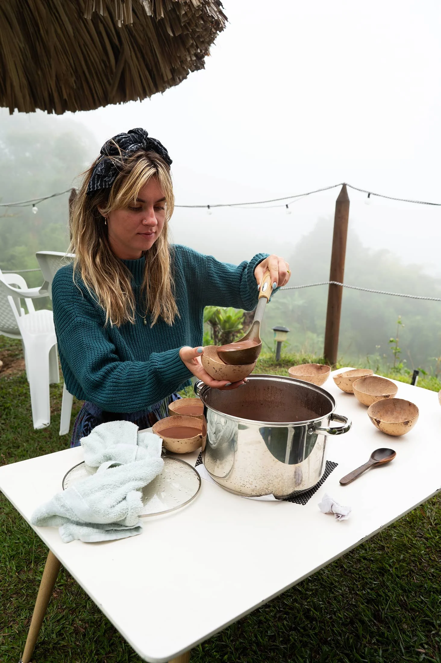 A woman with blonde hair, wearing a black bandana and blue sweater, is serving soup from a large pot into small bowls on an outdoor table. There are several bowls, a spoon, and a white towel on the table, with a foggy background, string lights, and greenery.