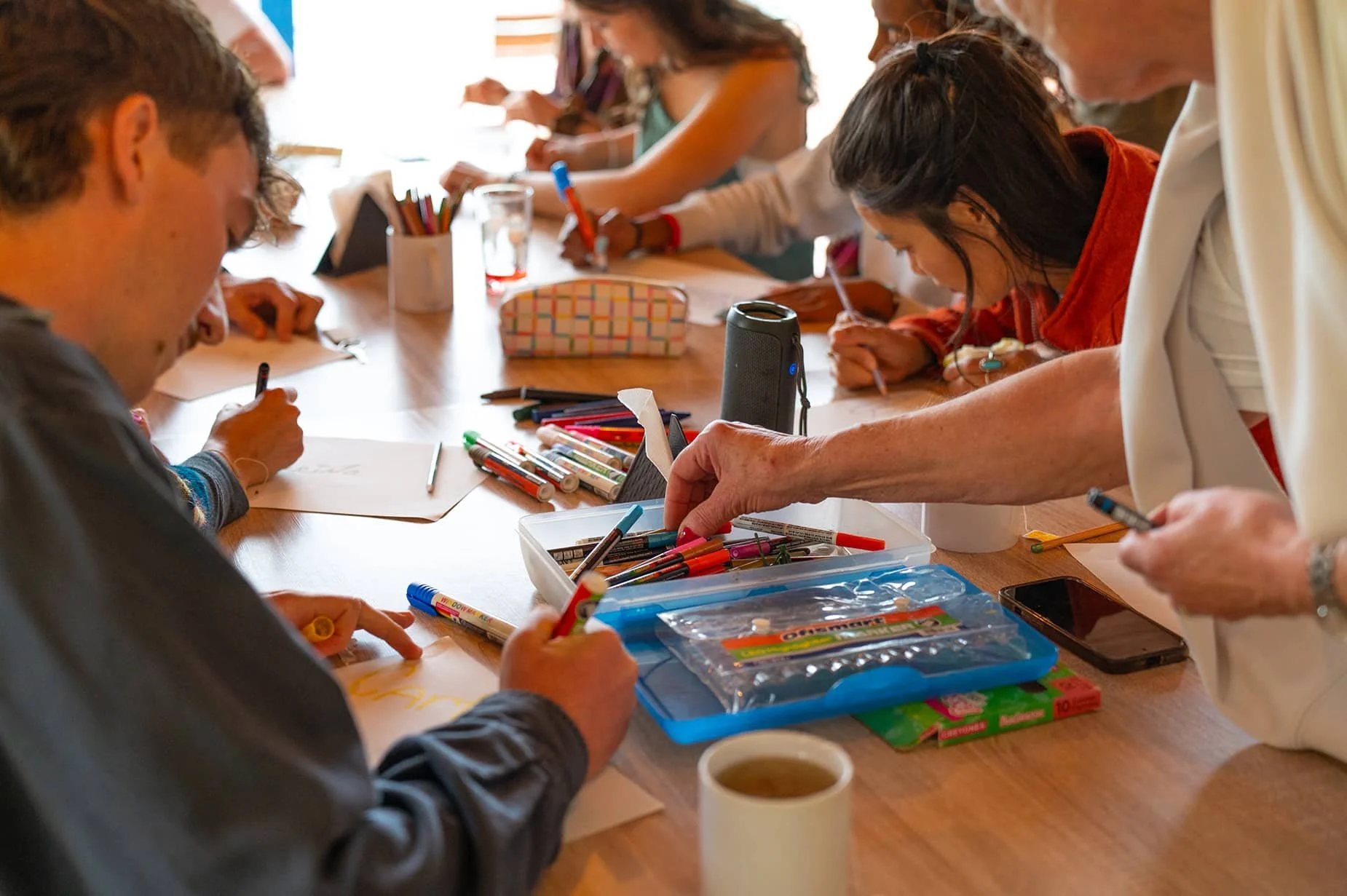 A group of people gather around a wooden table engaging in arts and crafts activities. They are using markers, colored pencils, and other supplies, with some writing and drawing on paper. There are various art materials and a mug on the table.
