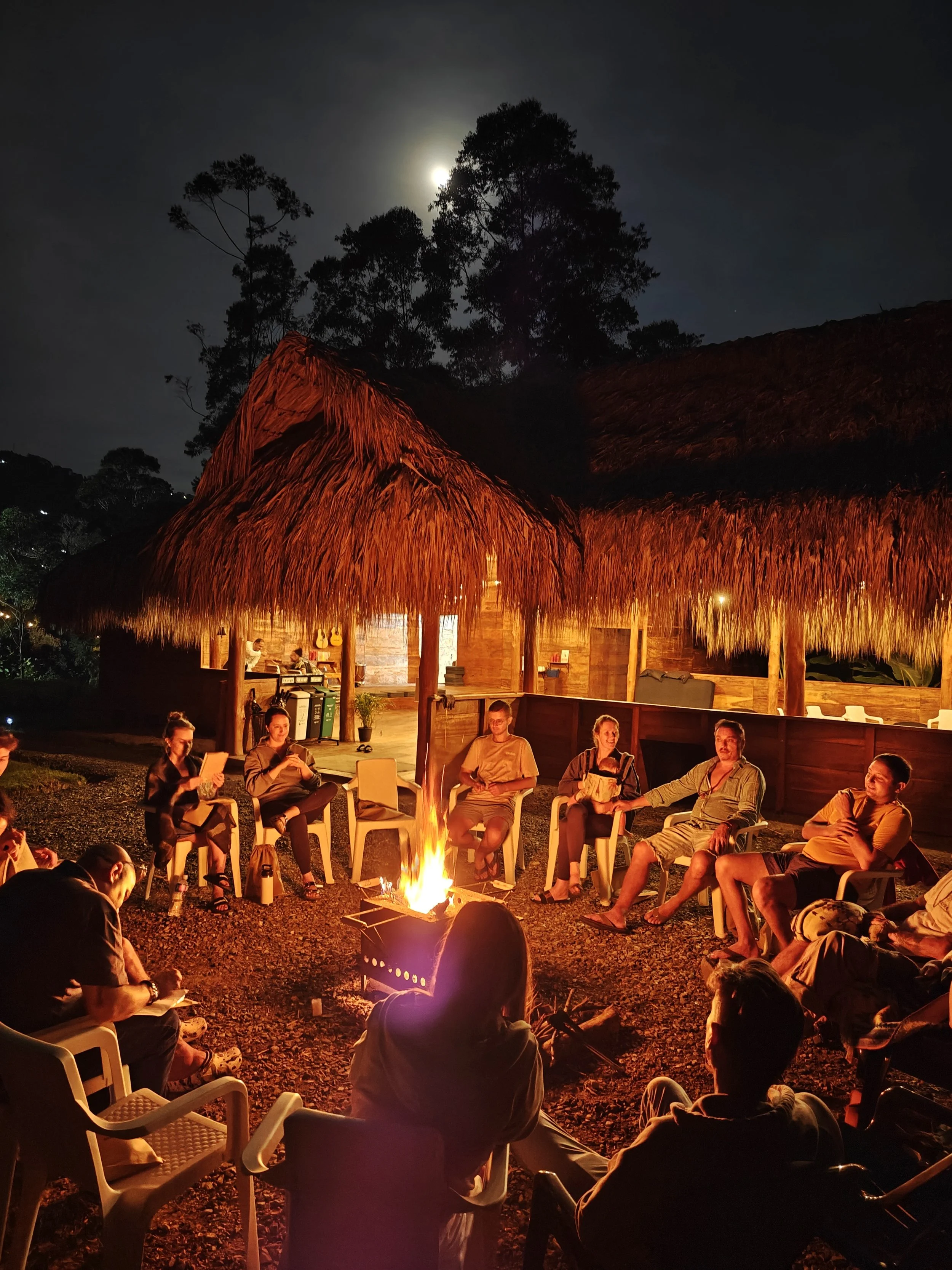 People sitting in chairs around a campfire at night, under a thatched roof structure with a full moon in the sky, surrounded by trees.
