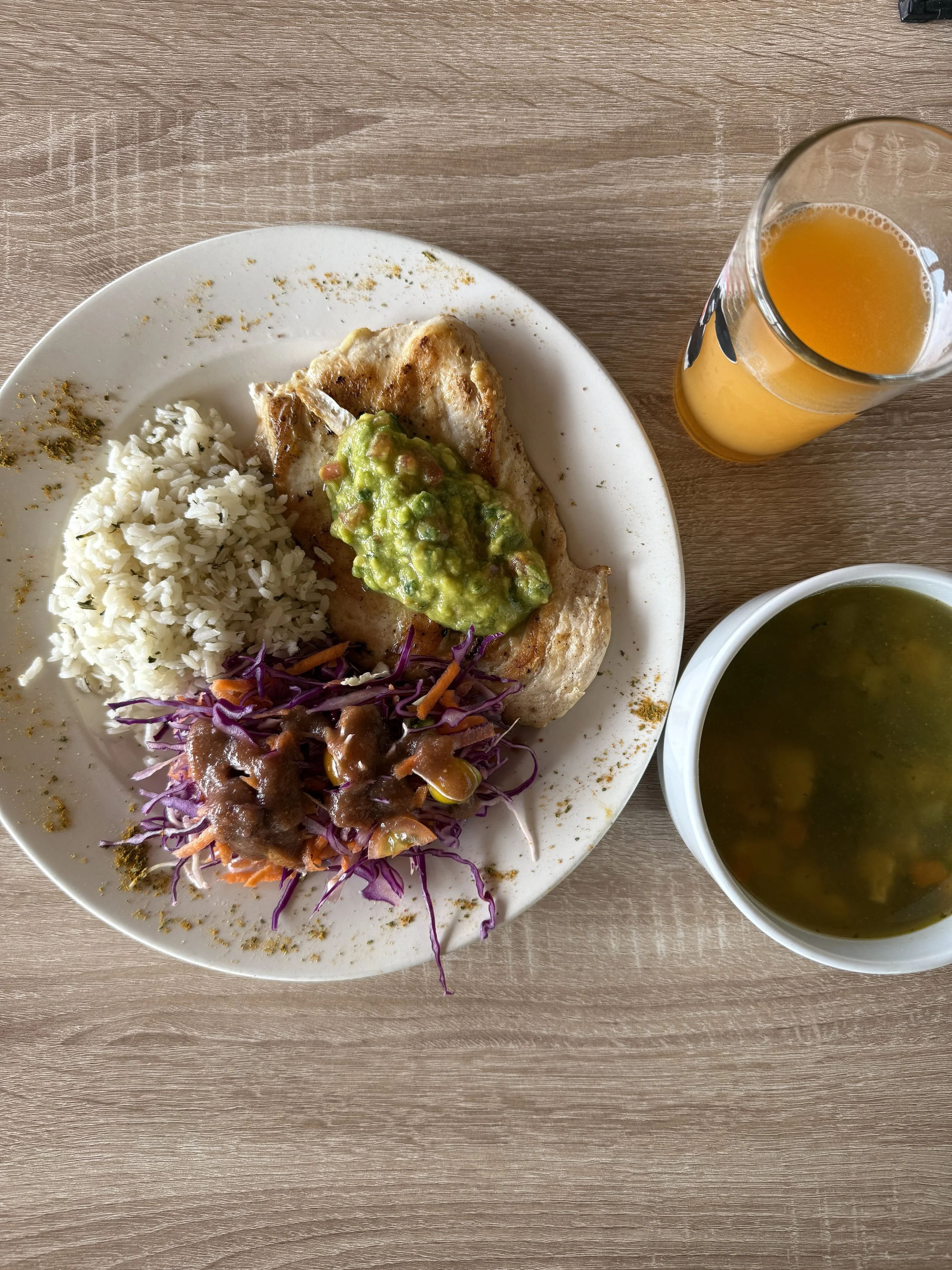 A plate of Mexican food with rice, grilled meat topped with guacamole, purple cabbage slaw, and a small serving of beans. Next to the plate are a glass of orange juice and a bowl of green soup, placed on a light brown wooden table.