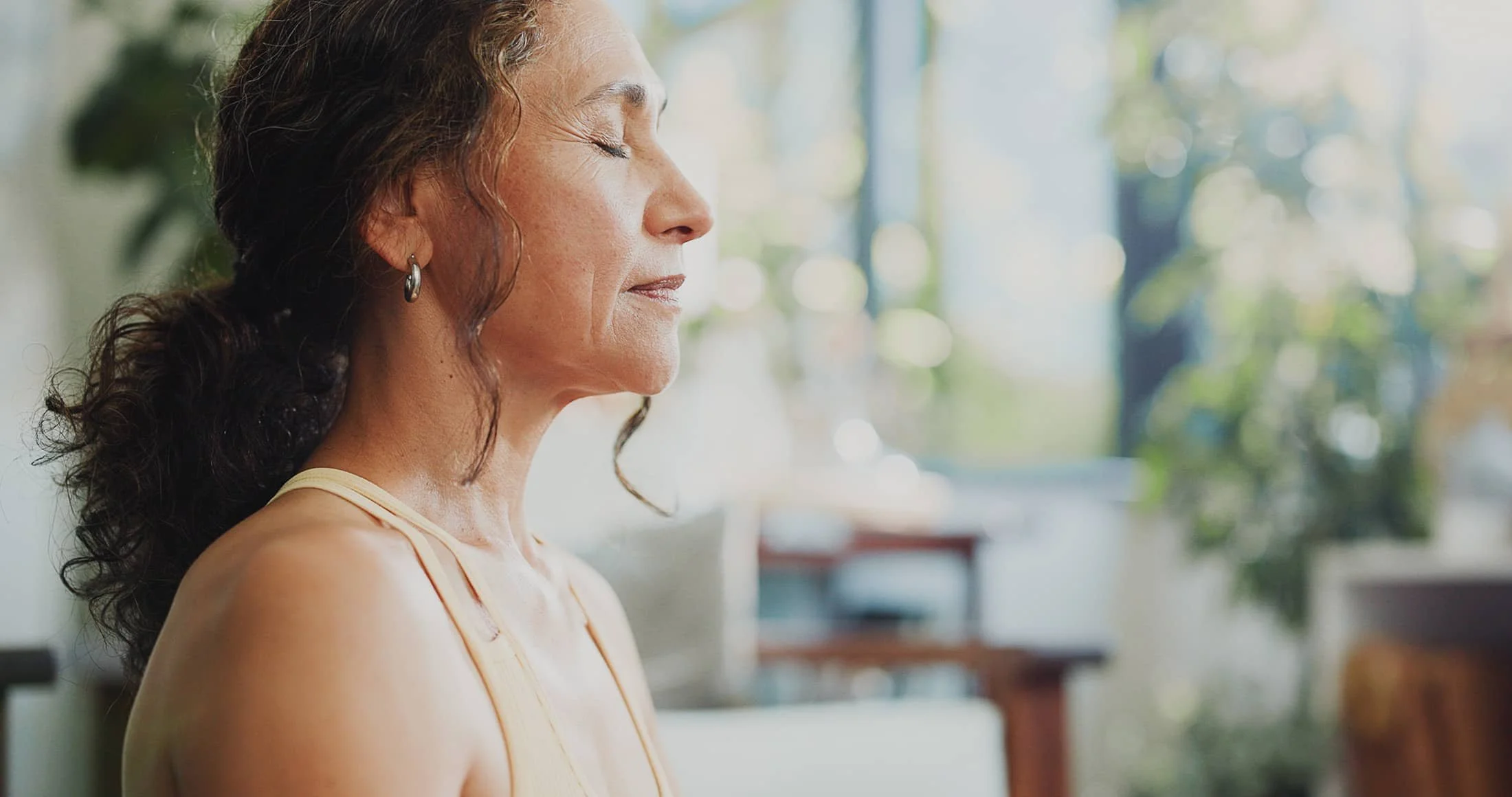 A woman with curly hair, earrings, and a sleeveless top sitting with her eyes closed in a bright, naturally lit room with greenery outside.
