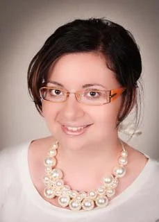 A woman with dark hair, wearing glasses, a white top, and a pearl necklace, smiling at the camera.